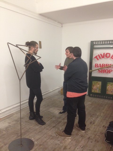 Three people are standing and talking indoors near a coat rack and a sign for a barbershop. One person is wearing glasses and has long hair, while the other two have short hair. The setting appears to be a casual or rehearsal space with wooden flooring.