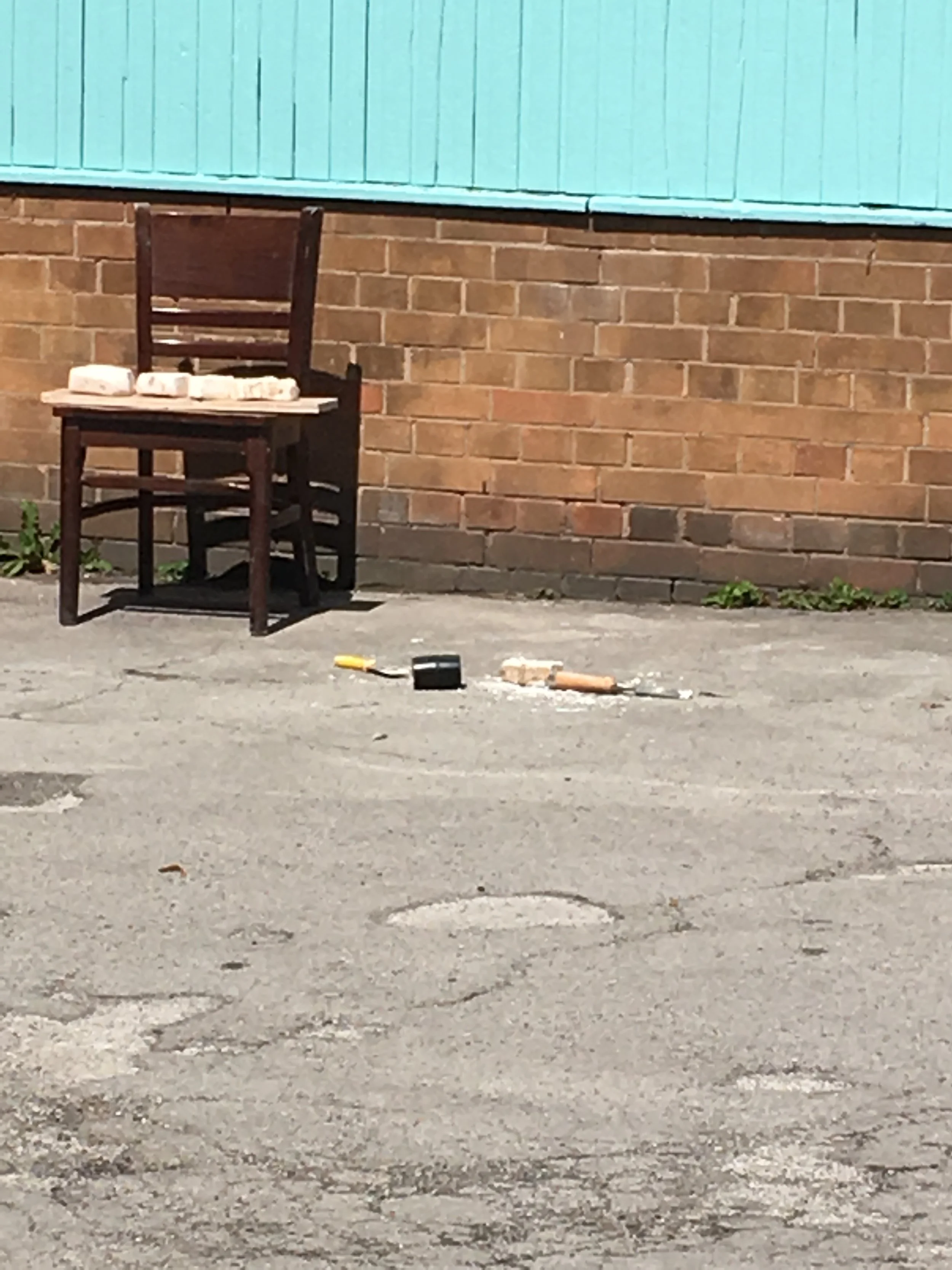 An outdoor scene with a brick wall and a teal wooden panel above it. There is a wooden chair with bricks on its seat, on a concrete or asphalt ground with some tools and debris scattered nearby.