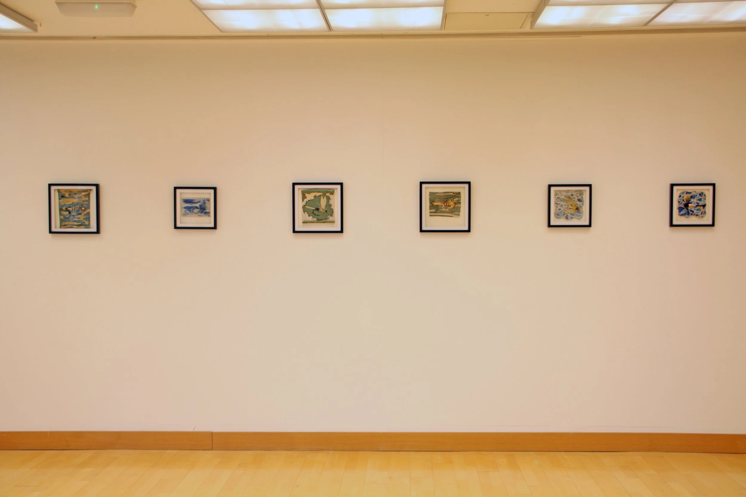 A gallery wall with six framed paintings in a museum or art gallery. The paintings are evenly spaced on a white wall. The room has a wooden floor and a ceiling with rectangular lights.