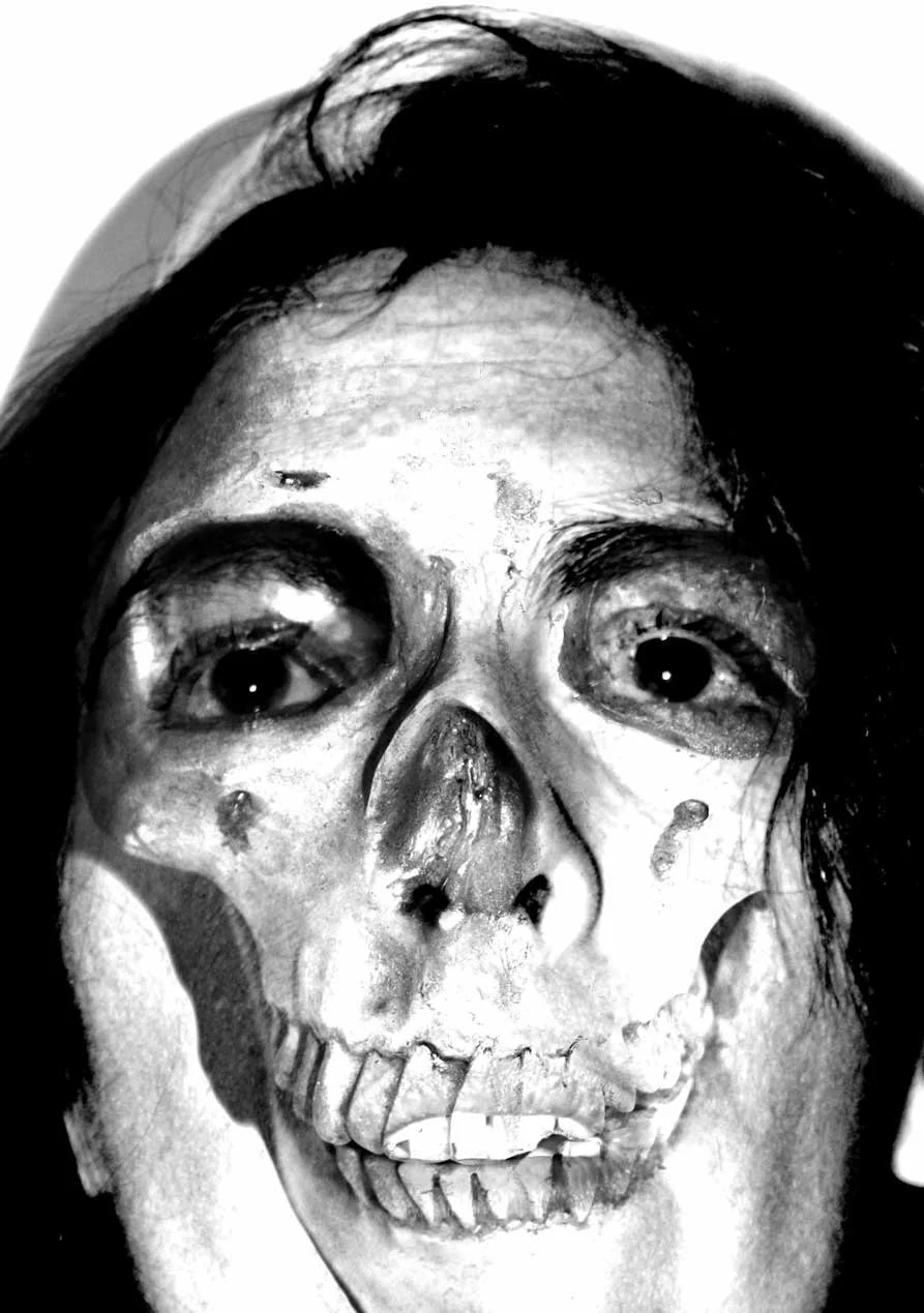 A black and white close-up photograph of a person's face partially covered with a skull overlay, giving a skeletal appearance.