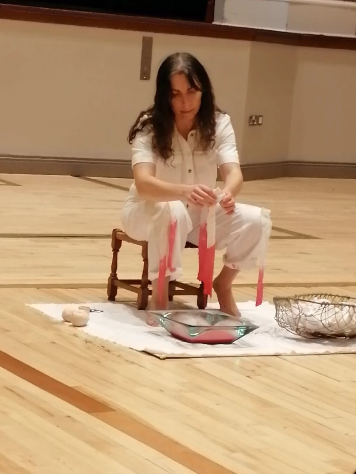 A woman with dark hair sits on a small wooden chair on a wooden floor, performing a ritual or ceremonial act with pink ribbons, a glass bowl, a white cloth, and various objects around her.
