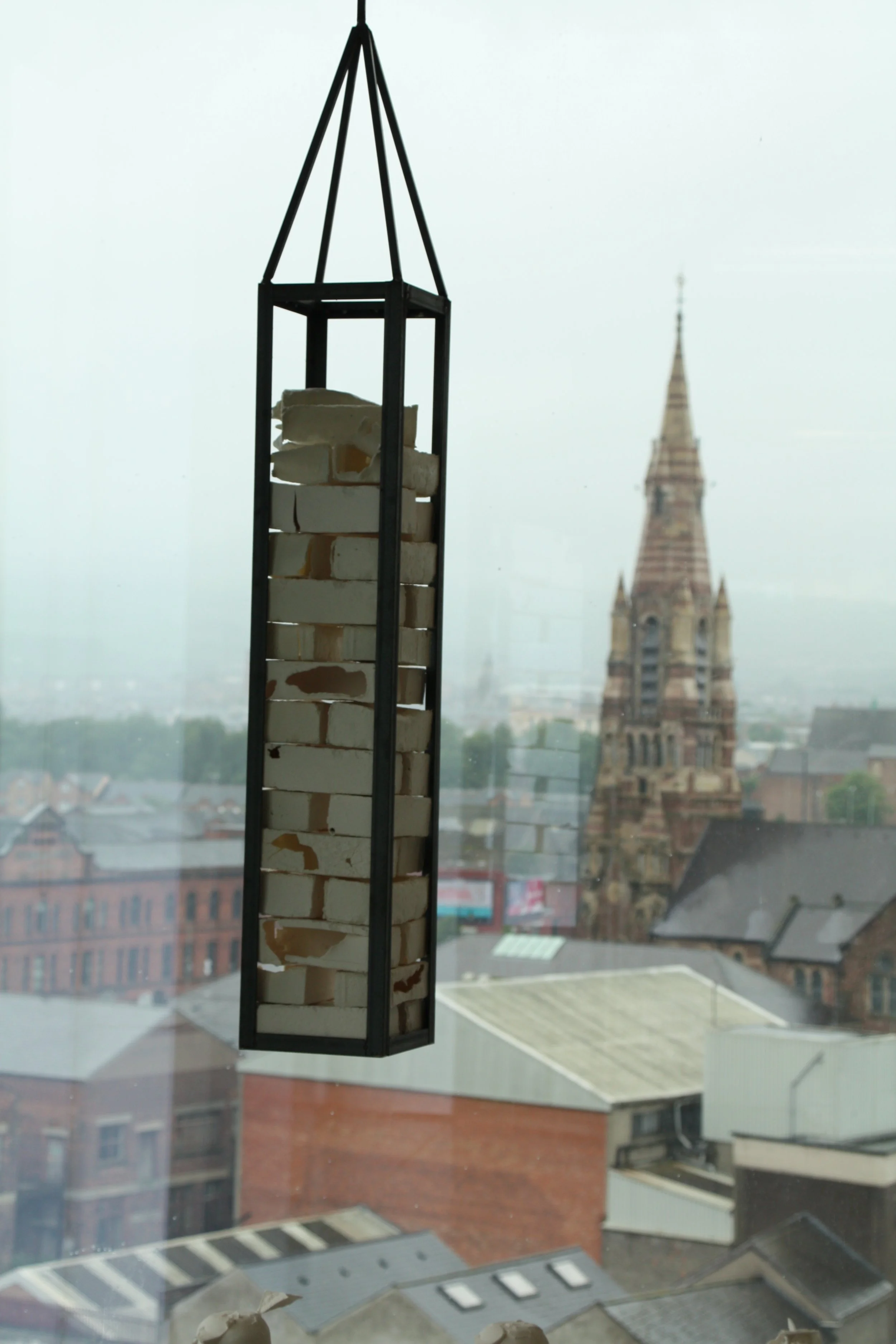 A window view of an urban cityscape with various buildings, including a church or cathedral with a tall, pointed spire, and a hanging decorative lantern with white bricks inside.