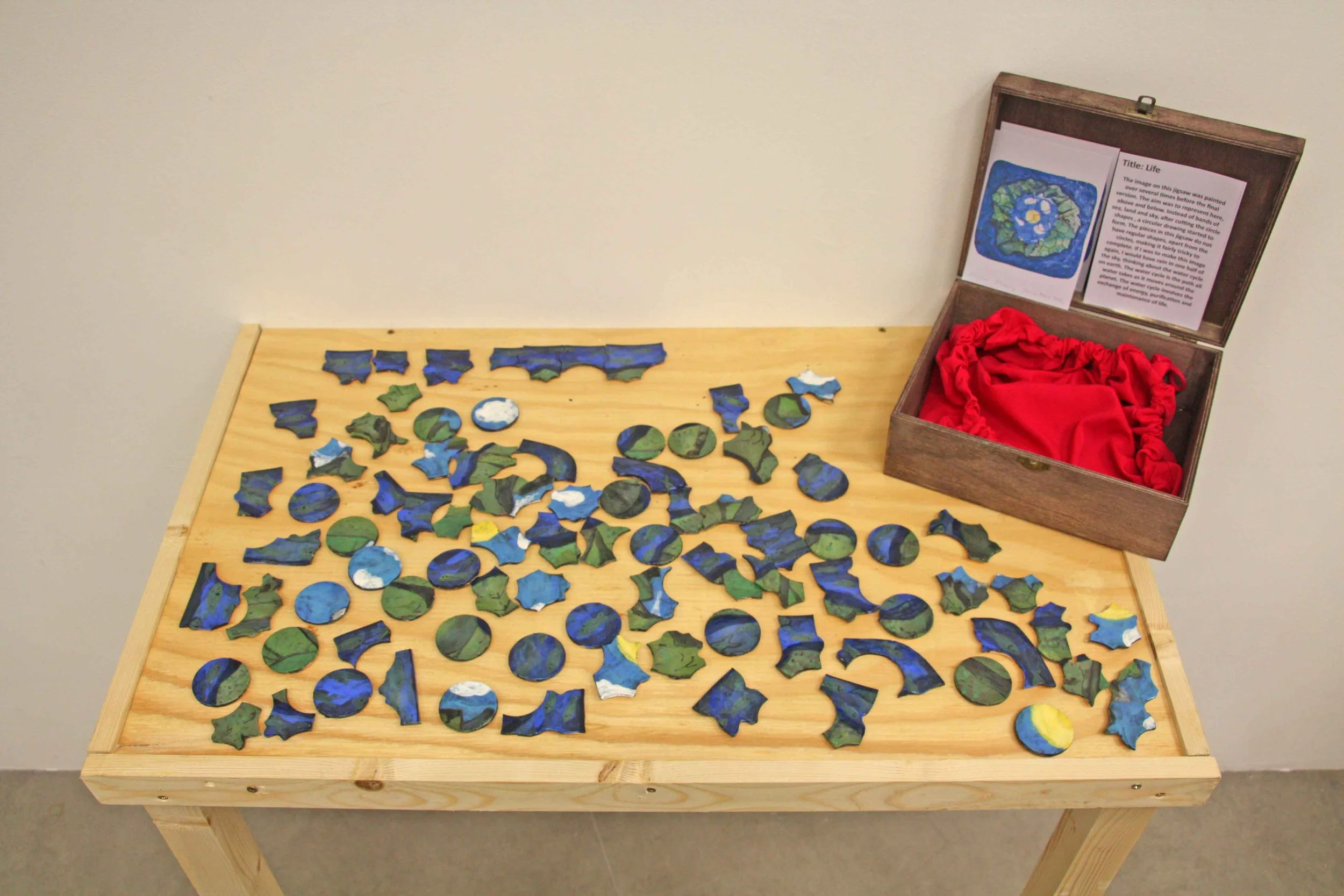 Colorful puzzle pieces scattered on a wooden table next to an open wooden box with a red cloth inside, containing a printed description of the puzzle titled 'Life' and its artwork.