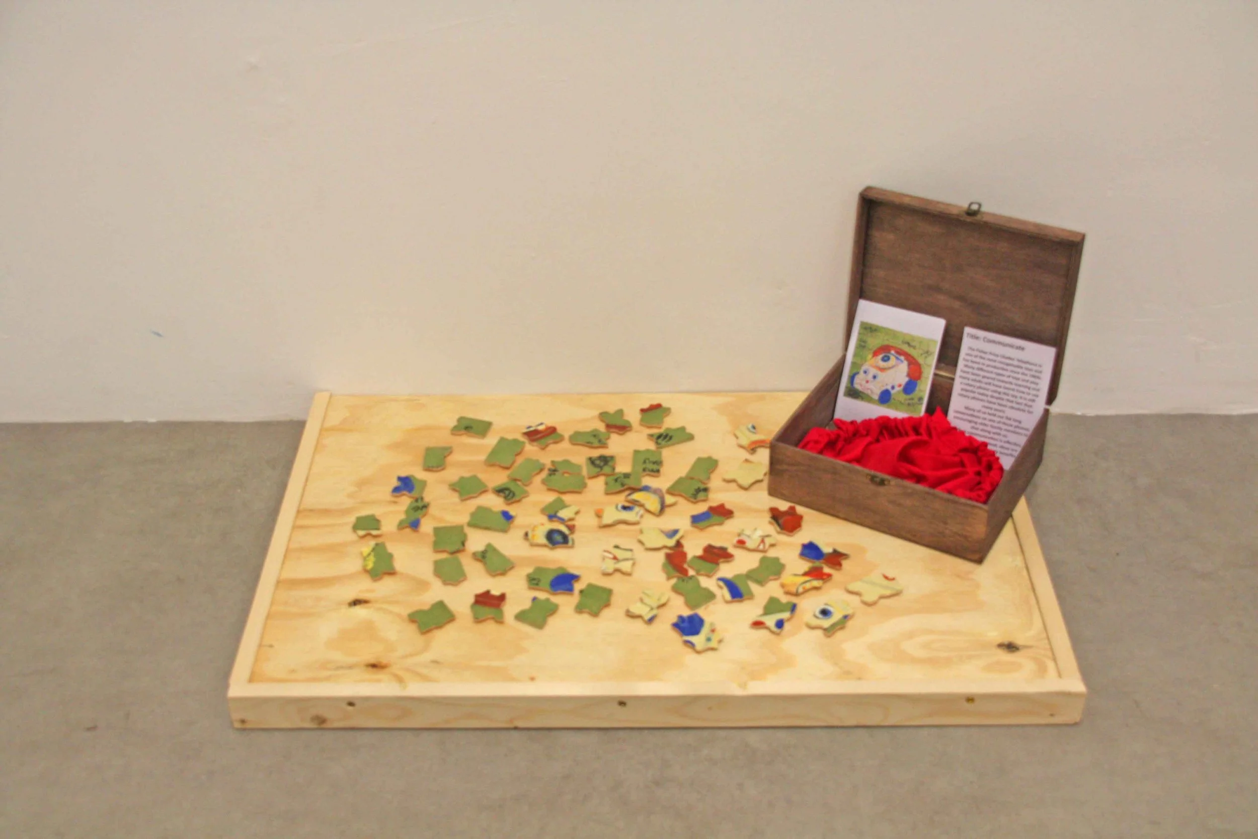 A wooden table with scattered puzzle pieces, a wooden box with red fabric lining, an informational card with a colorful illustration, and a plain beige wall in the background.
