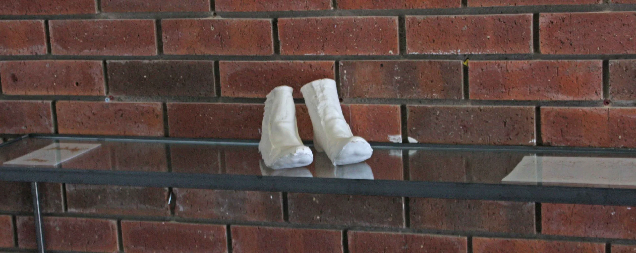 White plaster cast of a foot and ankle on a glass shelf against a red brick wall.