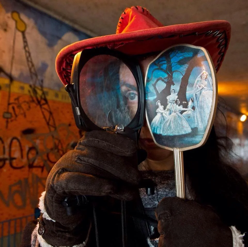 A person wearing a red cowboy hat and black gloves holds a fan with an image of ballet dancers in front of their face, creating a playful mask at a graffiti-covered location with warm lighting.