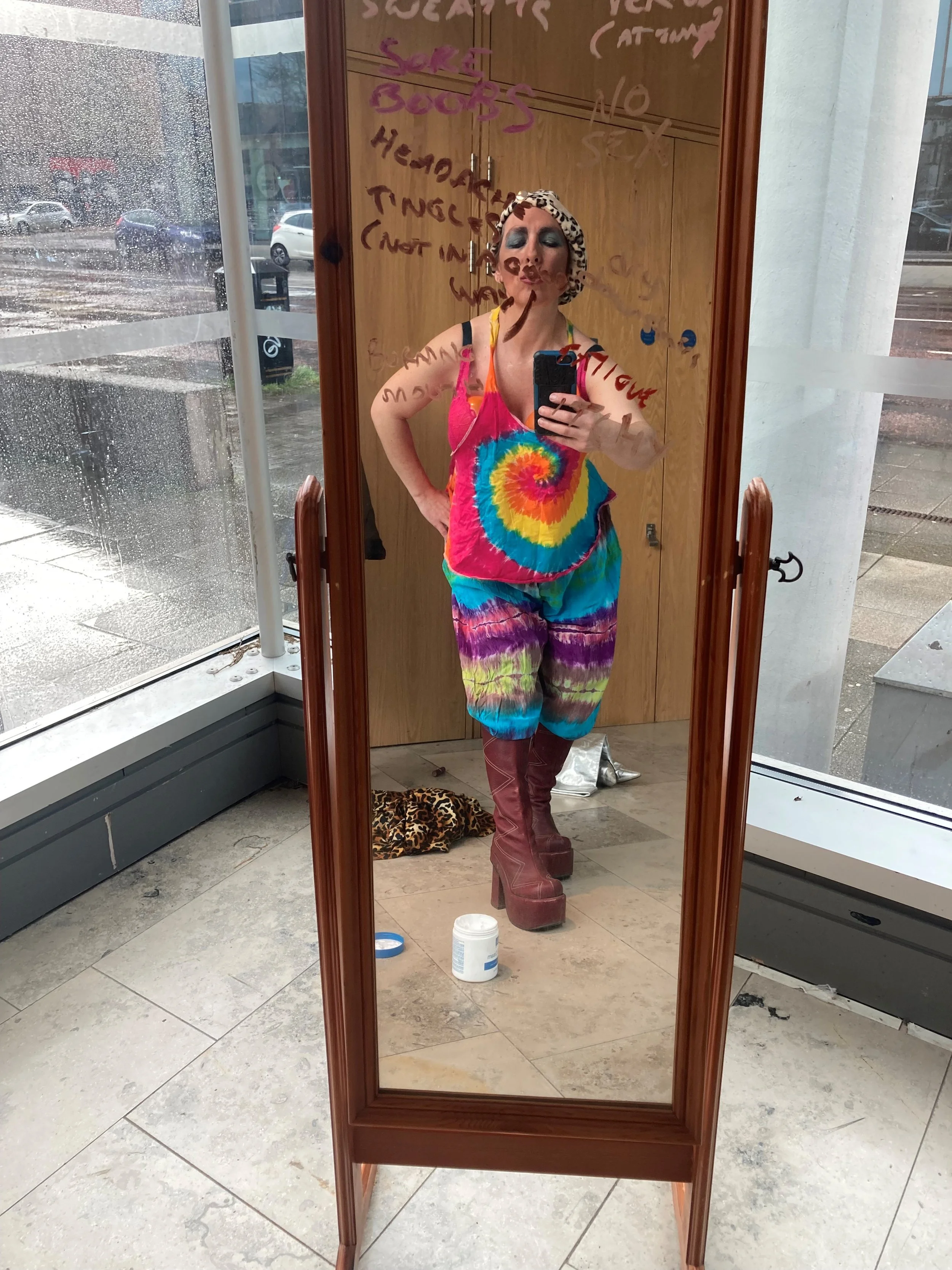 A woman taking a selfie in a mirror, wearing a colorful tie-dye outfit, tall platform boots, a leopard print headscarf, and bold makeup. The mirror has handwritten notes on it, and there is a leopard-print cloth on the floor. Outside the window, rain