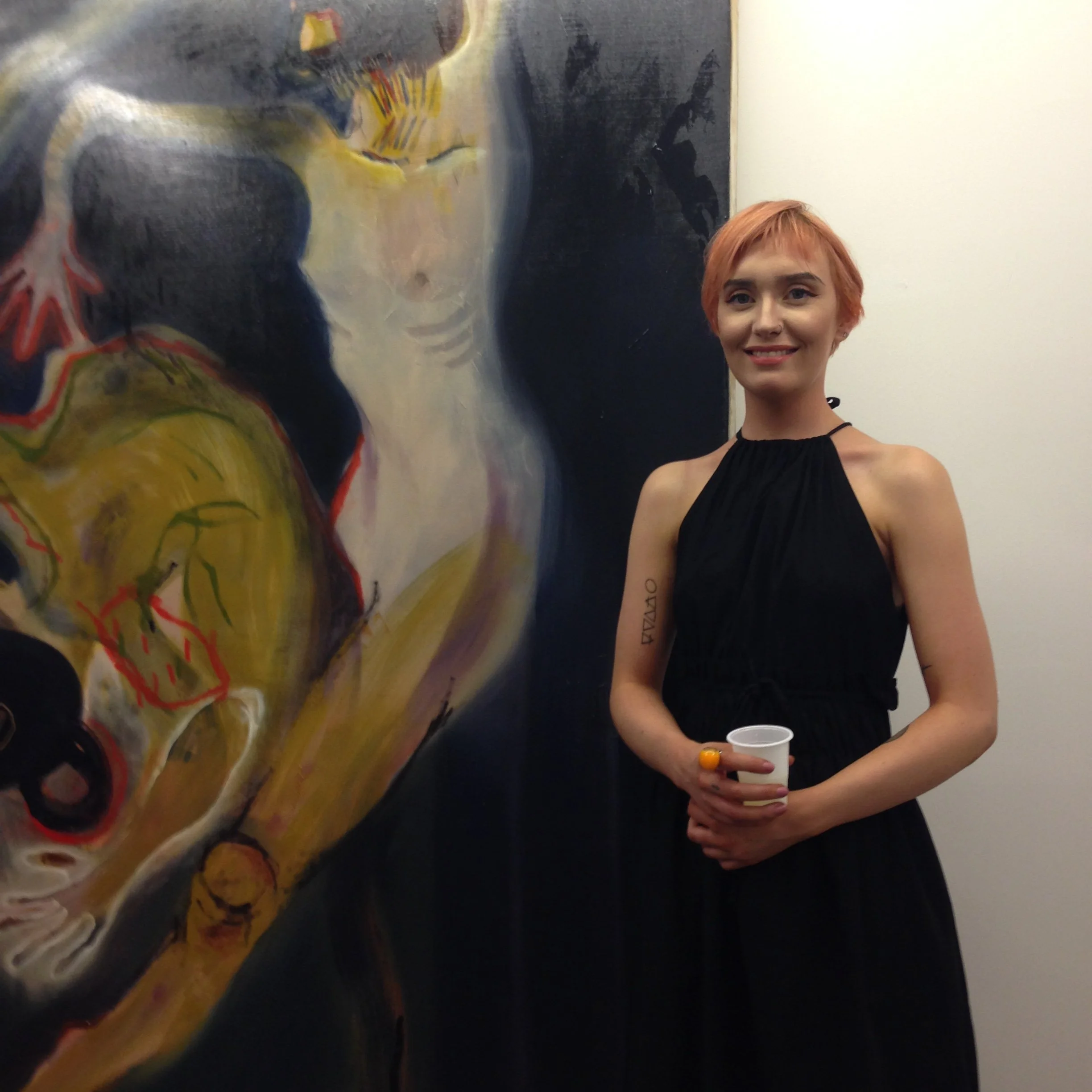 Young woman with short red hair smiling, wearing a black top with a tattoo on her right forearm, holding a cup, standing next to abstract art painting on a white wall.