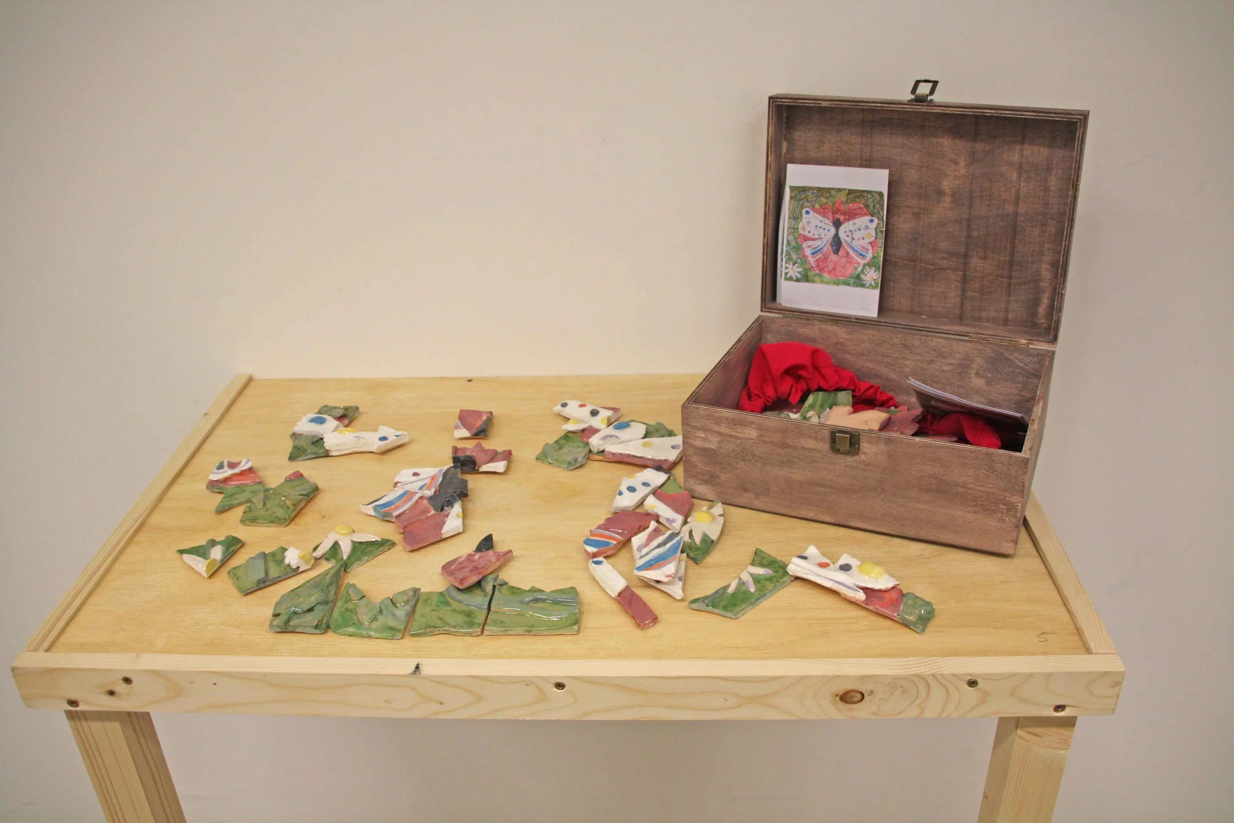 A wooden table displaying colorful broken pottery jigsaw pieces across its surface. An open wooden box with a butterfly drawing on the inside lid sits on the table, containing red fabric and some pottery pieces. The background is plain and off-white.