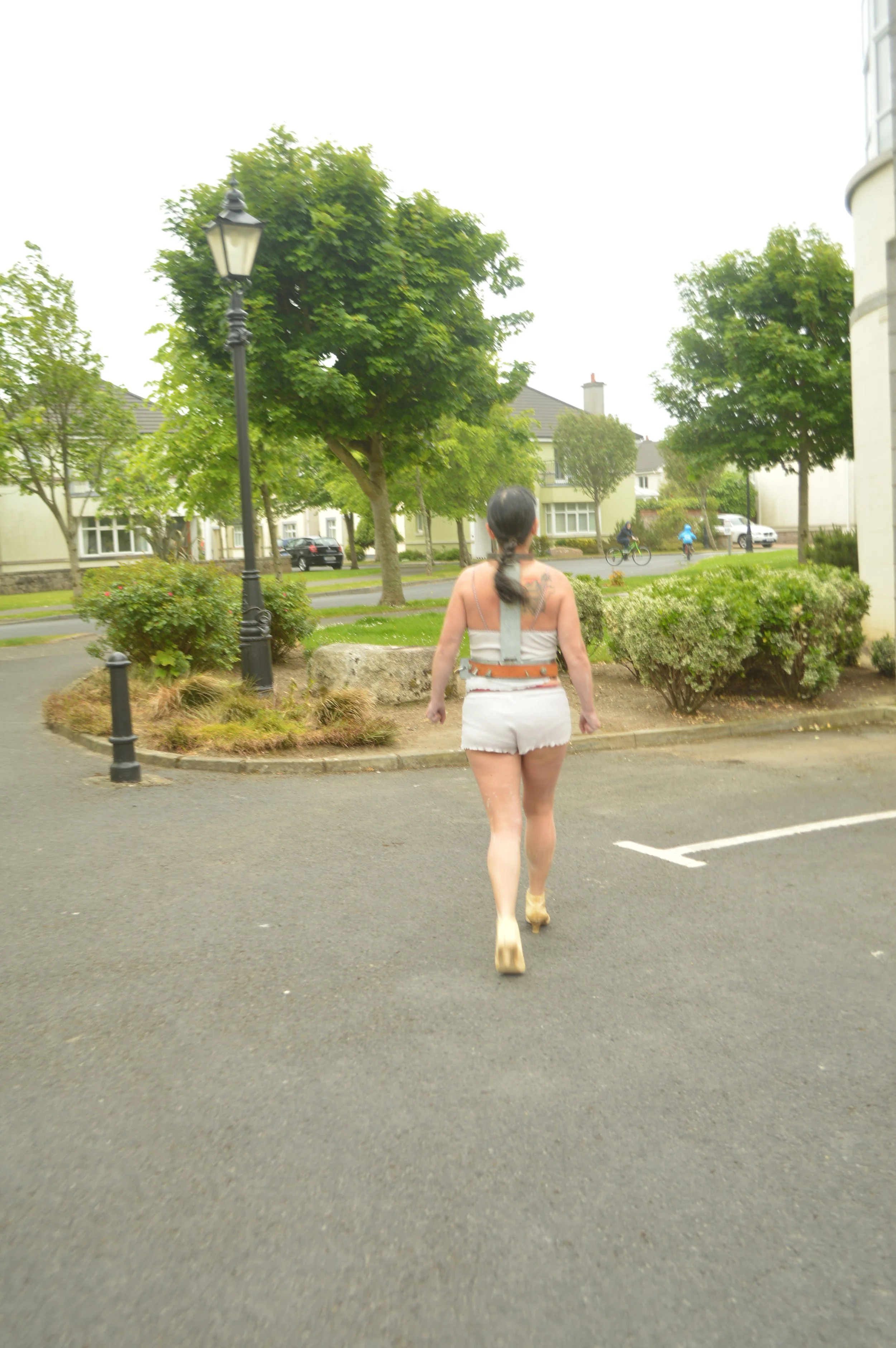Woman with dark hair in ponytail wearing white outfit walking away in a parking lot, with trees and houses in the background.