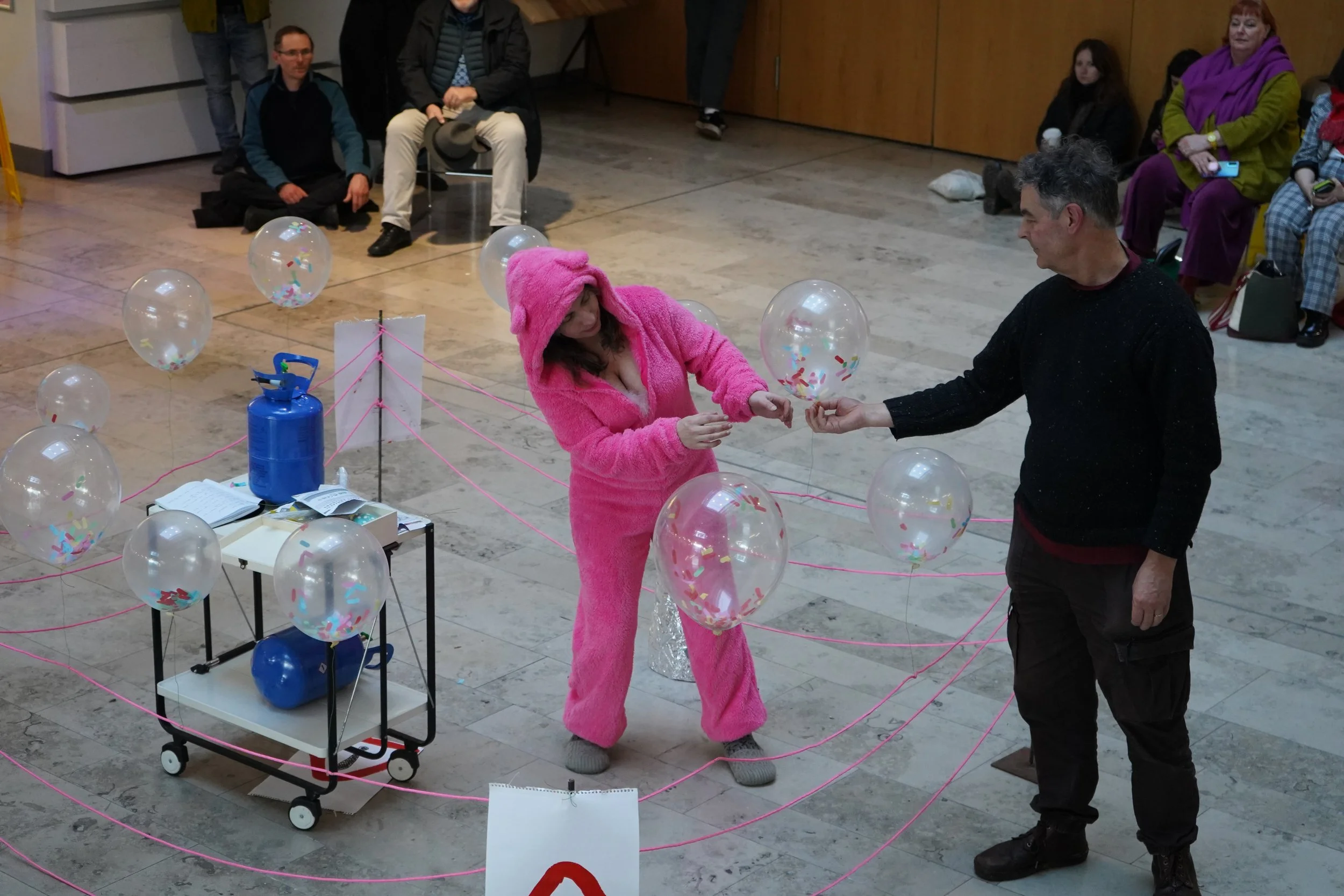 A woman dressed in a pink furry costume with a hood is handing something to a man in a black sweater, standing inside a maze marked by pink string ties. The maze contains transparent balloons with confetti inside, and there is a blue gas cylinder on 