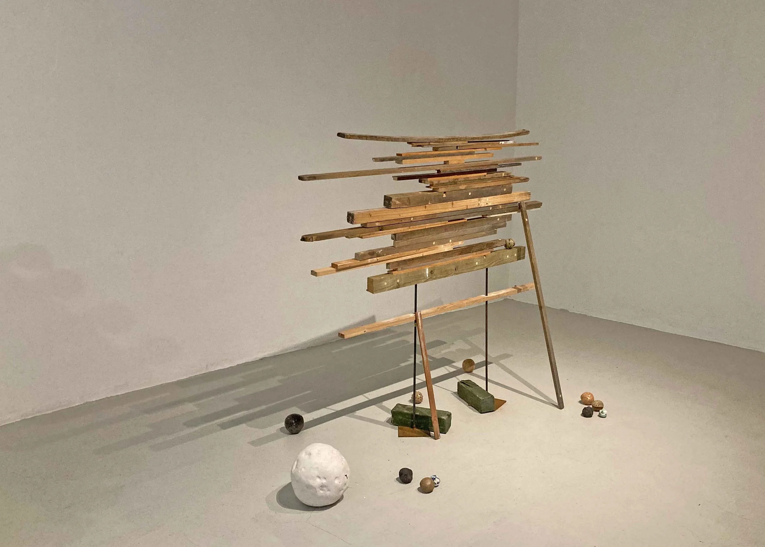 A wooden sculpture resembling a mountain landscape with horizontal wooden planks supported by a wooden frame, placed in a gallery with plain walls and scattered small spherical objects on the floor.