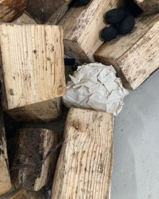 Wooden logs stacked with a crumpled white paper ceramic object  and a small cluster of coal on top
