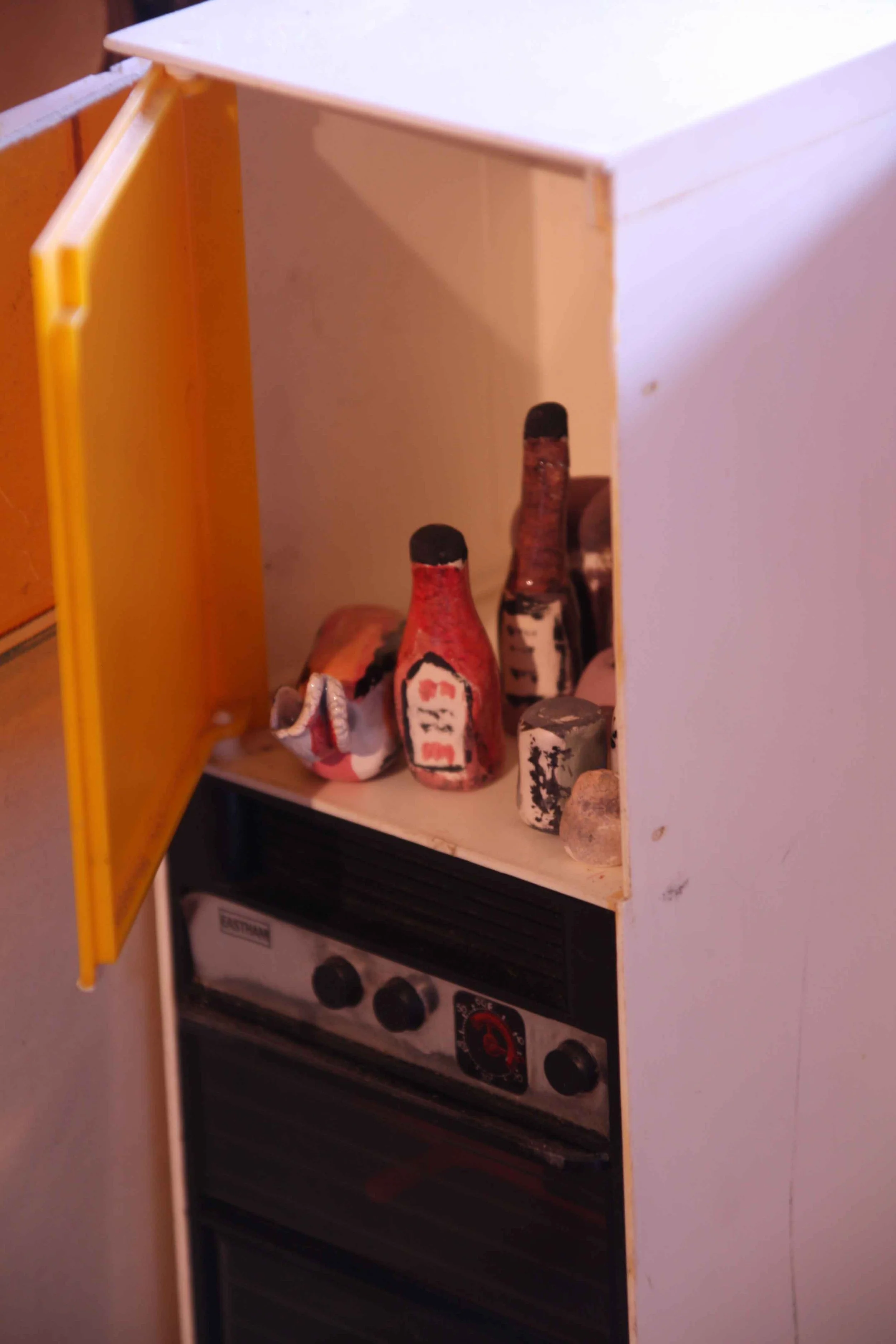 Small toy stove with a white cabinet and yellow door, shelf with toy bottles and cans, oven with black knobs.