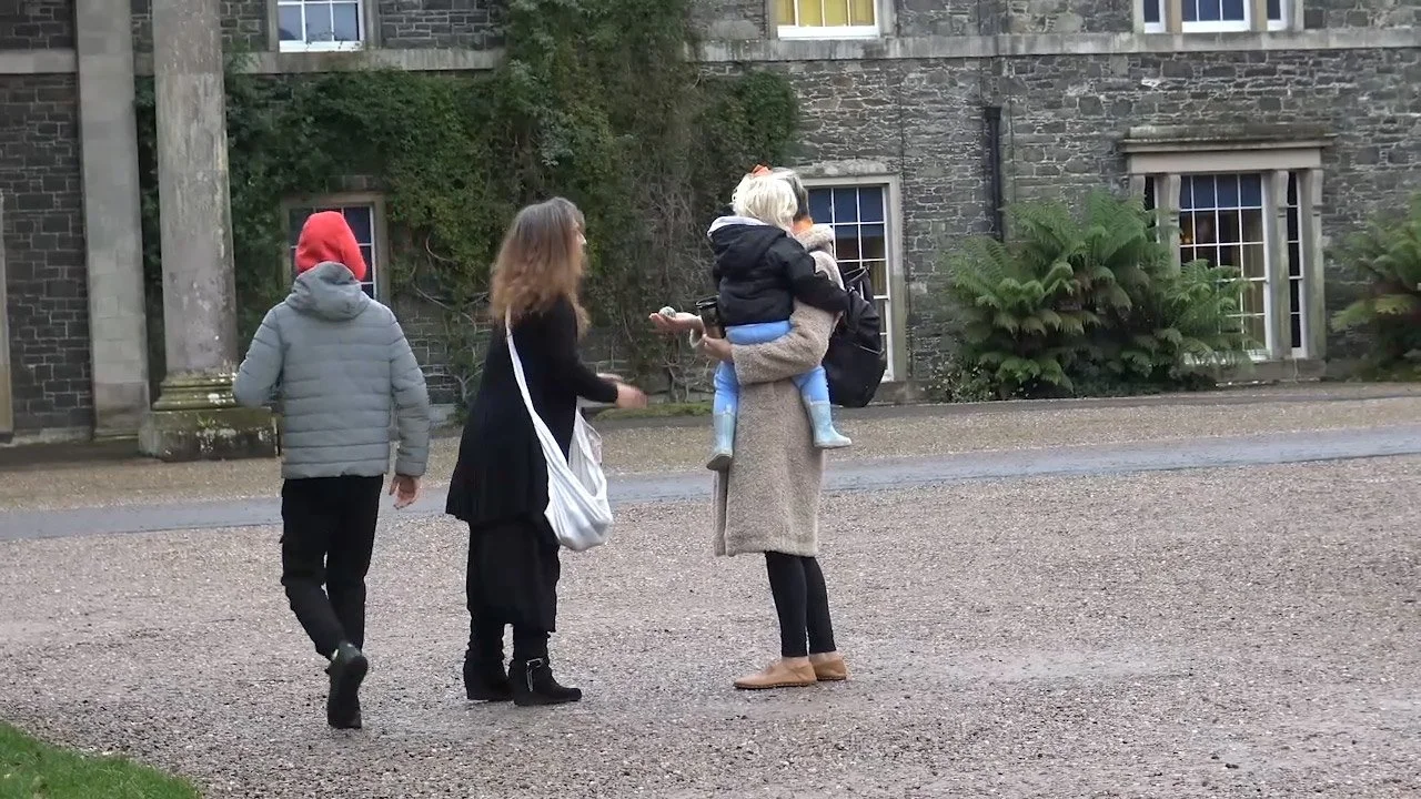 Four people walking outdoors in front of a gray stone building with windows; one woman carries a child, others are walking nearby.