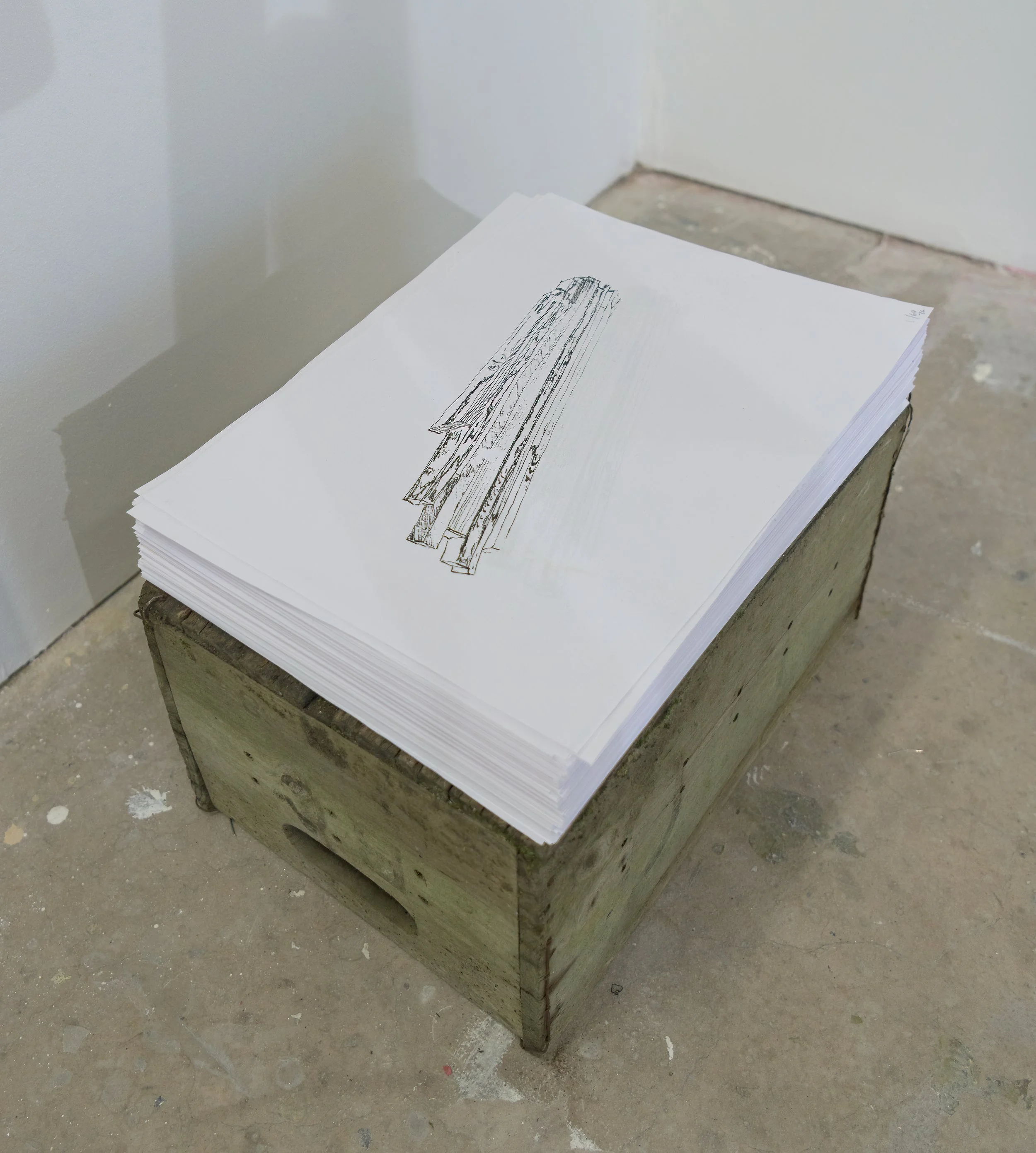 Stacks of white paper with black sketch drawings set on a rough wooden table in a room with concrete floor.