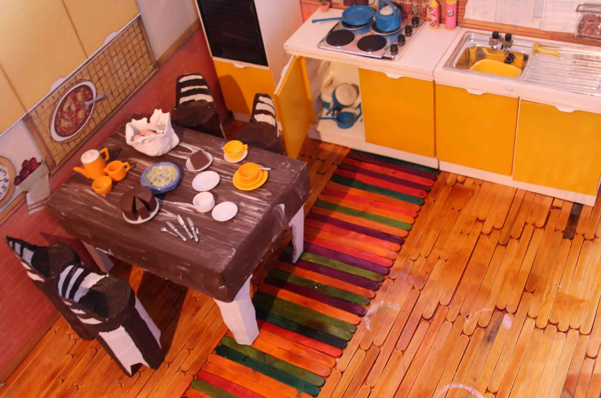 A miniature kitchen with yellow cabinets, a small stovetop, a sink, and a dining table with dishes and cups, all in a dollhouse setting.