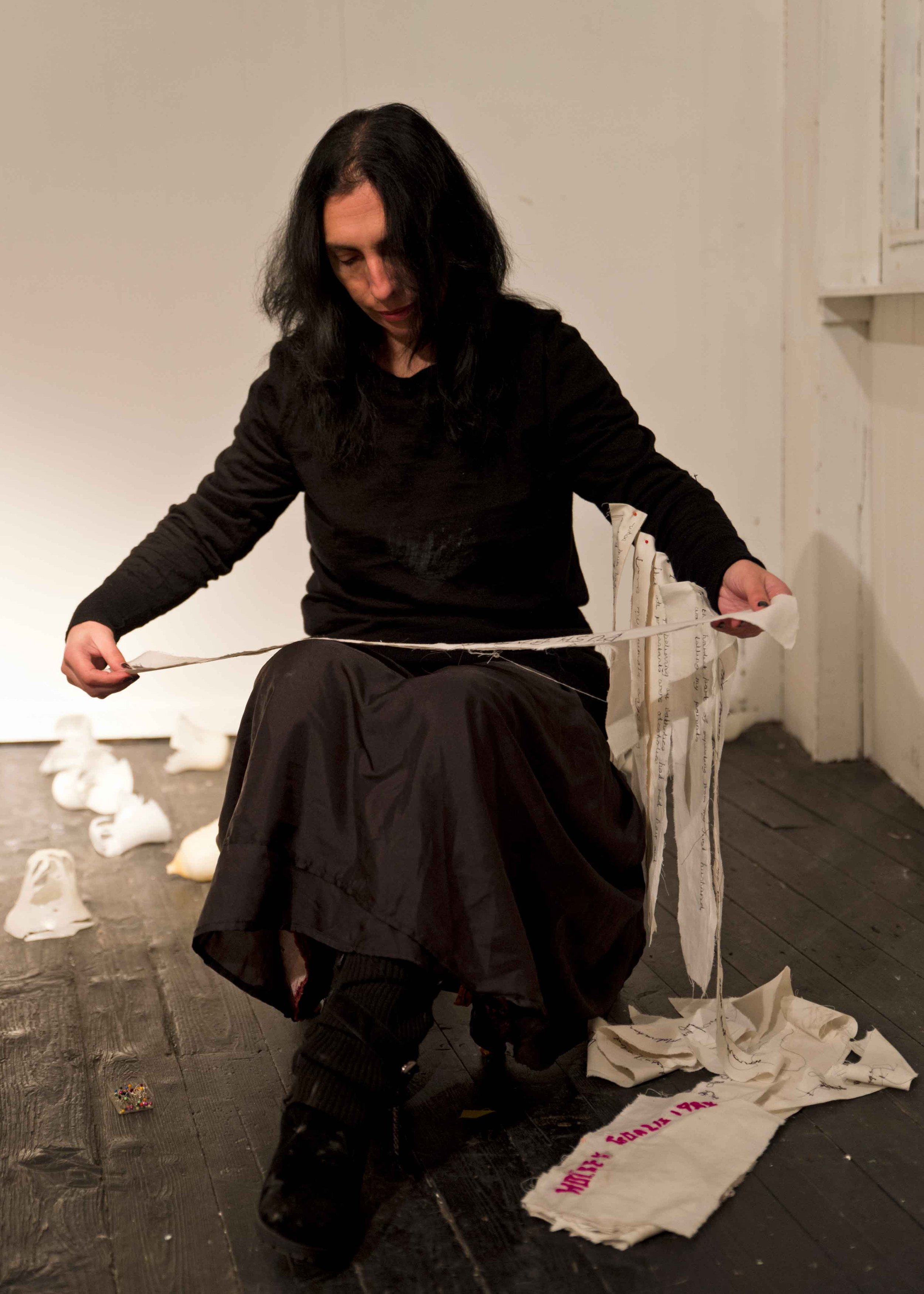 A woman with black hair wearing a black shirt and long skirt is sitting on a wooden floor, looking down while holding a torn piece of. Crumpled papers and a torn banner with pink writing are scattered around her. The background features a plain white