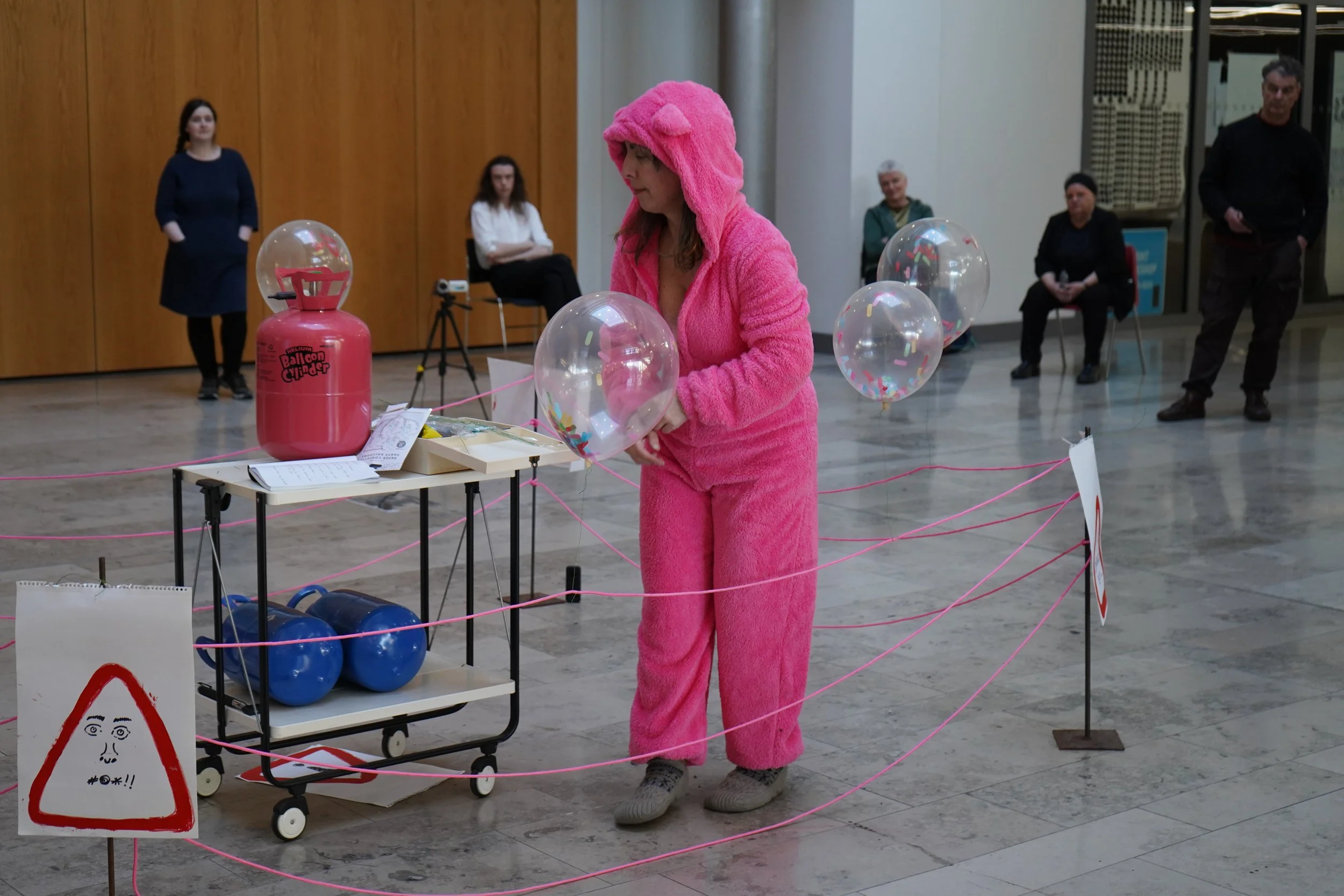 A woman dressed in a pink fuzzy onesie that resembles a bear costume, standing near balloons and a propane tank labeled 'Balloon Gunfire,' at an indoor event. Several people sit and observe in the background.