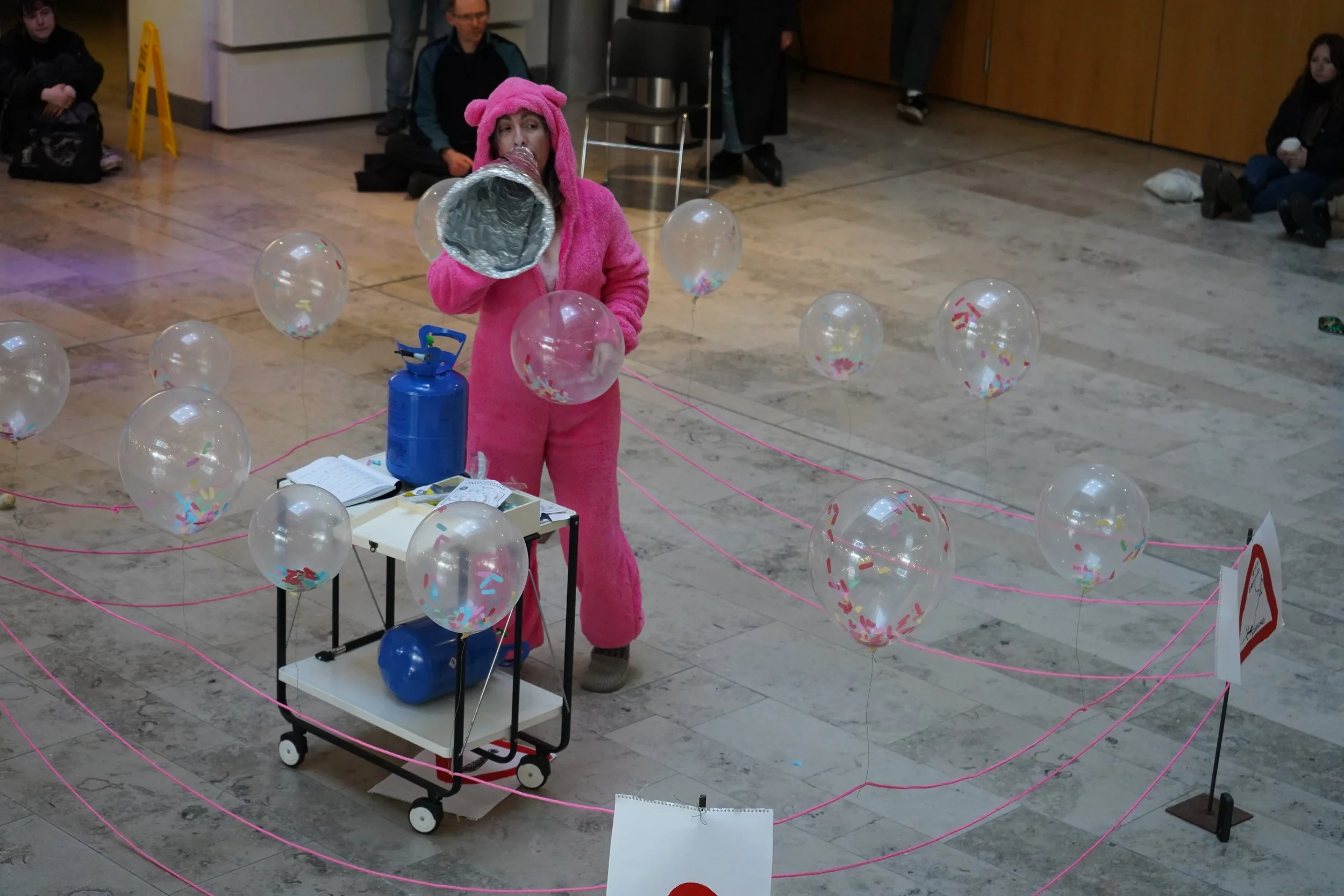 A woman in a pink bear costume blowing up balloons inside a public space with people seated along the walls, pink string lines on the floor creating a geometric pattern.