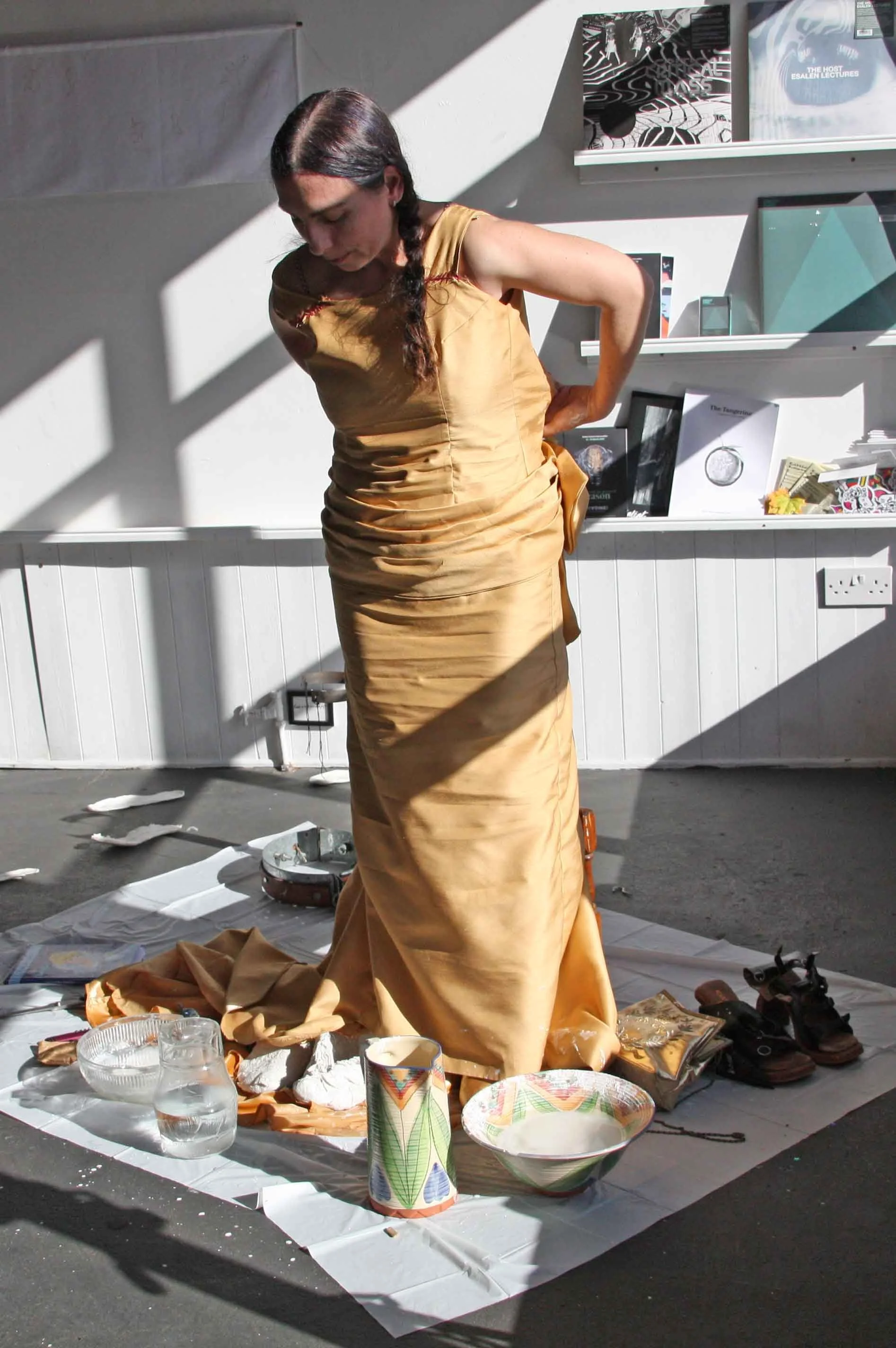 A woman in a regal gold satin dress with a side bow is standing with her hand on her back, amid a collection of dishes, cups, and shoes on the ground, in a space with white shelves displaying books and artwork, and bright sunlight casting shadows.
