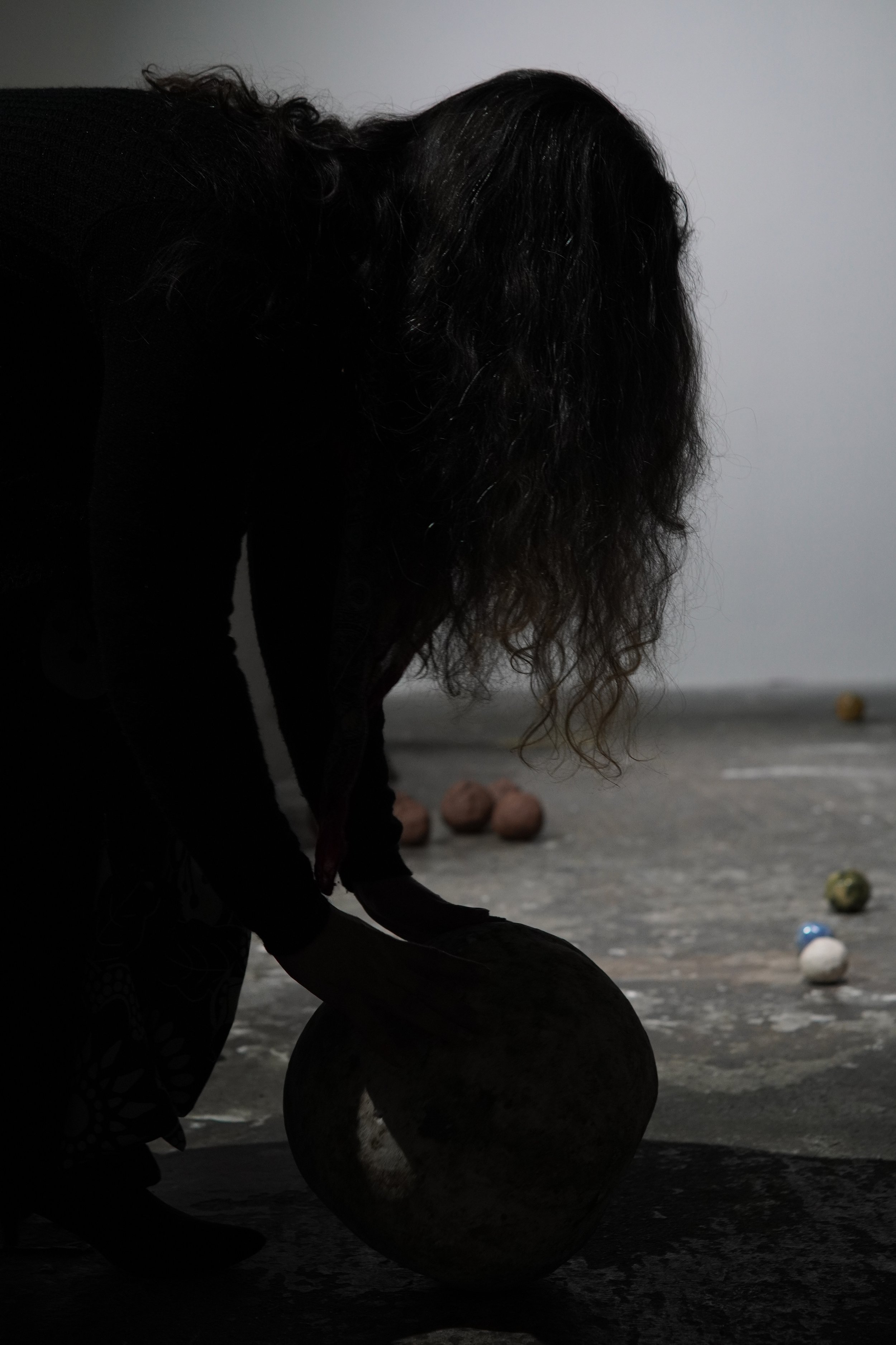 A person with long curly hair is kneeling and stacking colored stones on the ground, with a plain wall background.