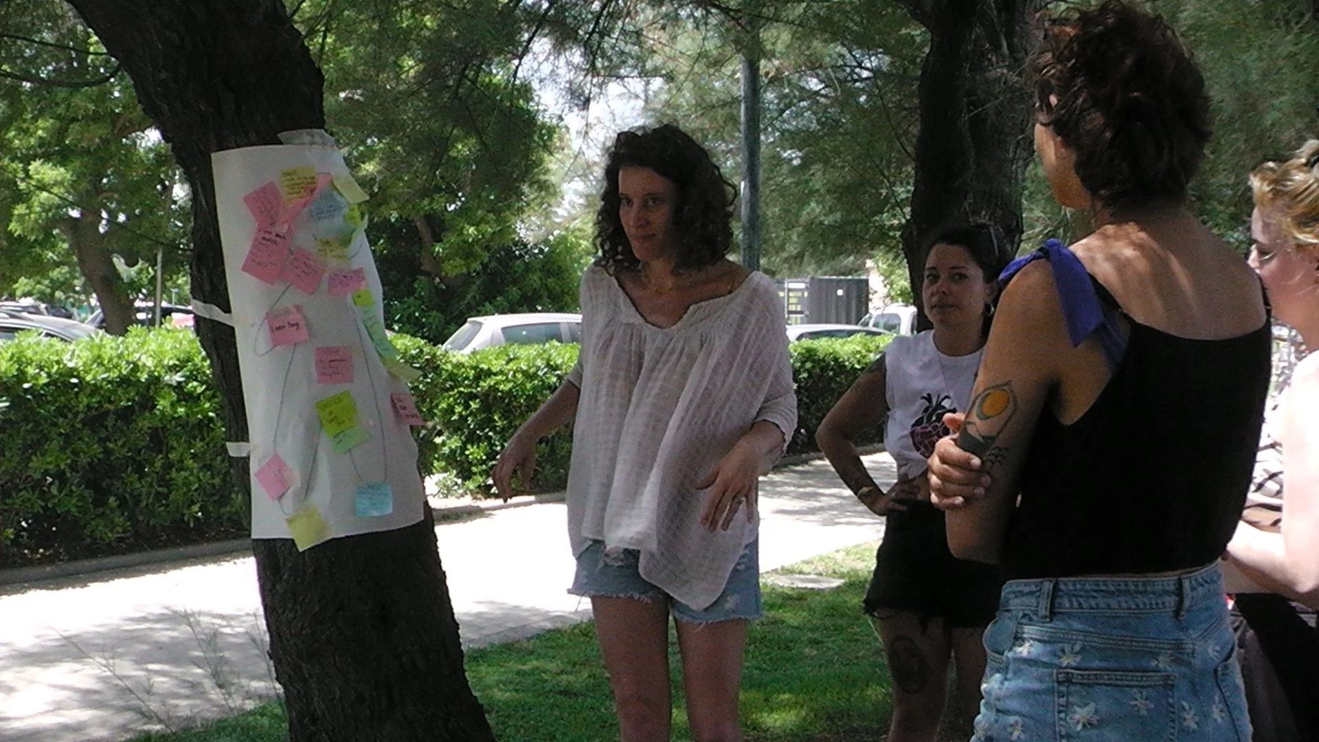 A group of four women standing outdoors under trees, engaged in a discussion around a large sheet of paper with colorful sticky notes attached, in a park or garden setting with parked cars in the background.