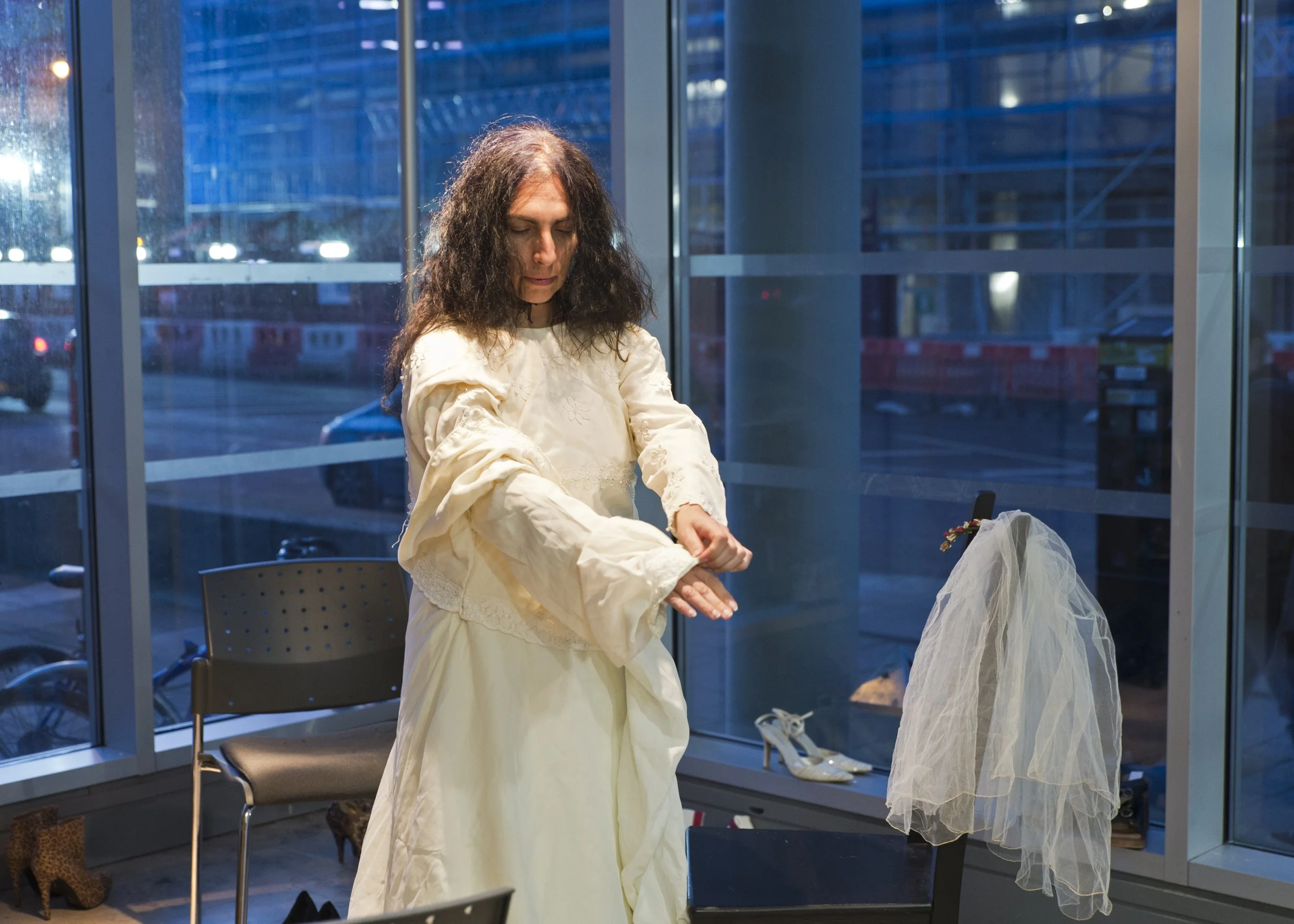 A woman with curly hair wearing a cream-colored dress is sitting in a modern, well-lit room with large windows at night. There are shoes and accessories on the floor near her, and a chair is behind her.