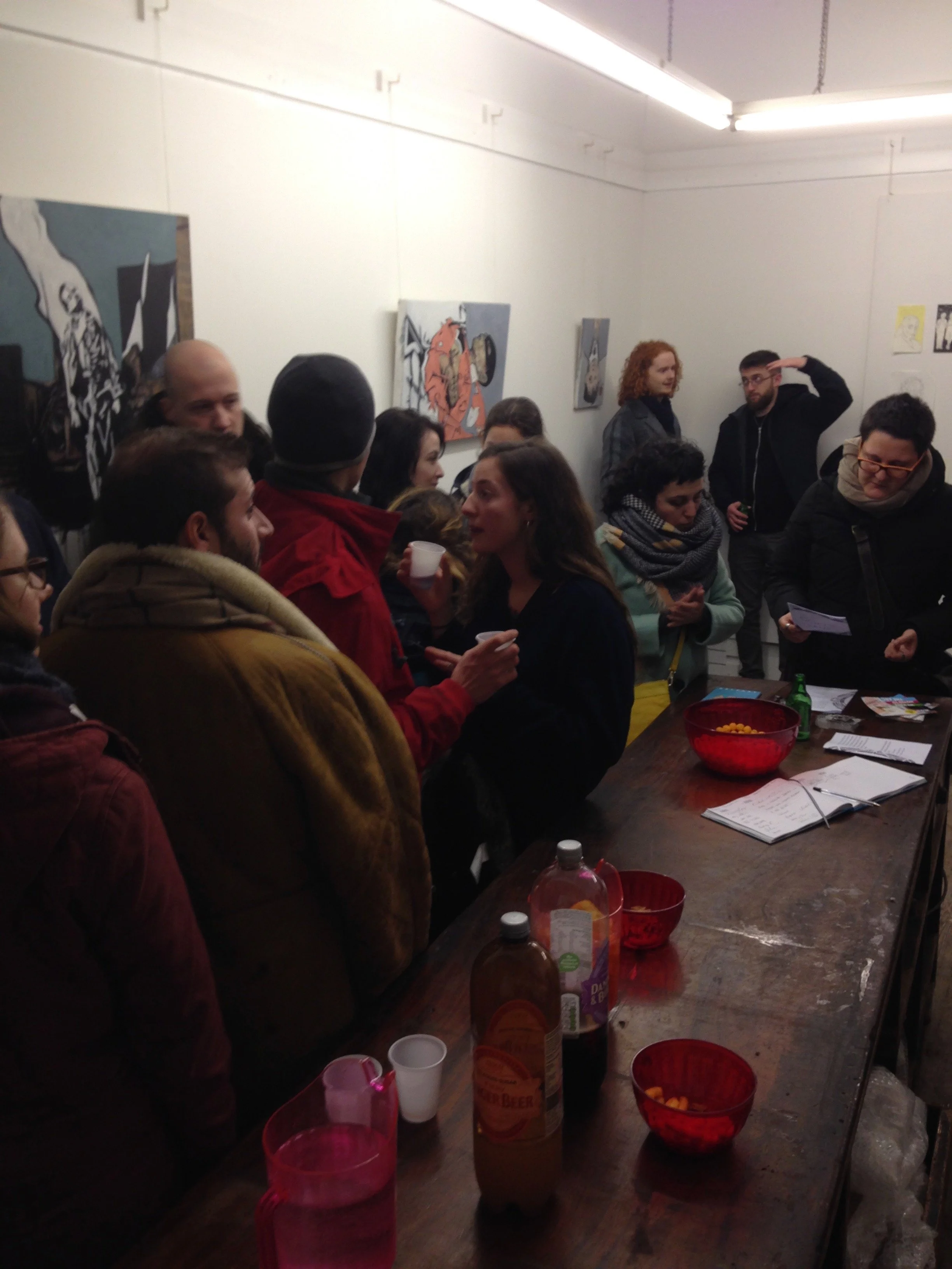 A group of people gathered in a small room, engaging in conversation. The room has white walls decorated with colorful artwork. On a table, there are cups, bottles, bowls with snacks, and papers, suggesting a casual social event or gathering.