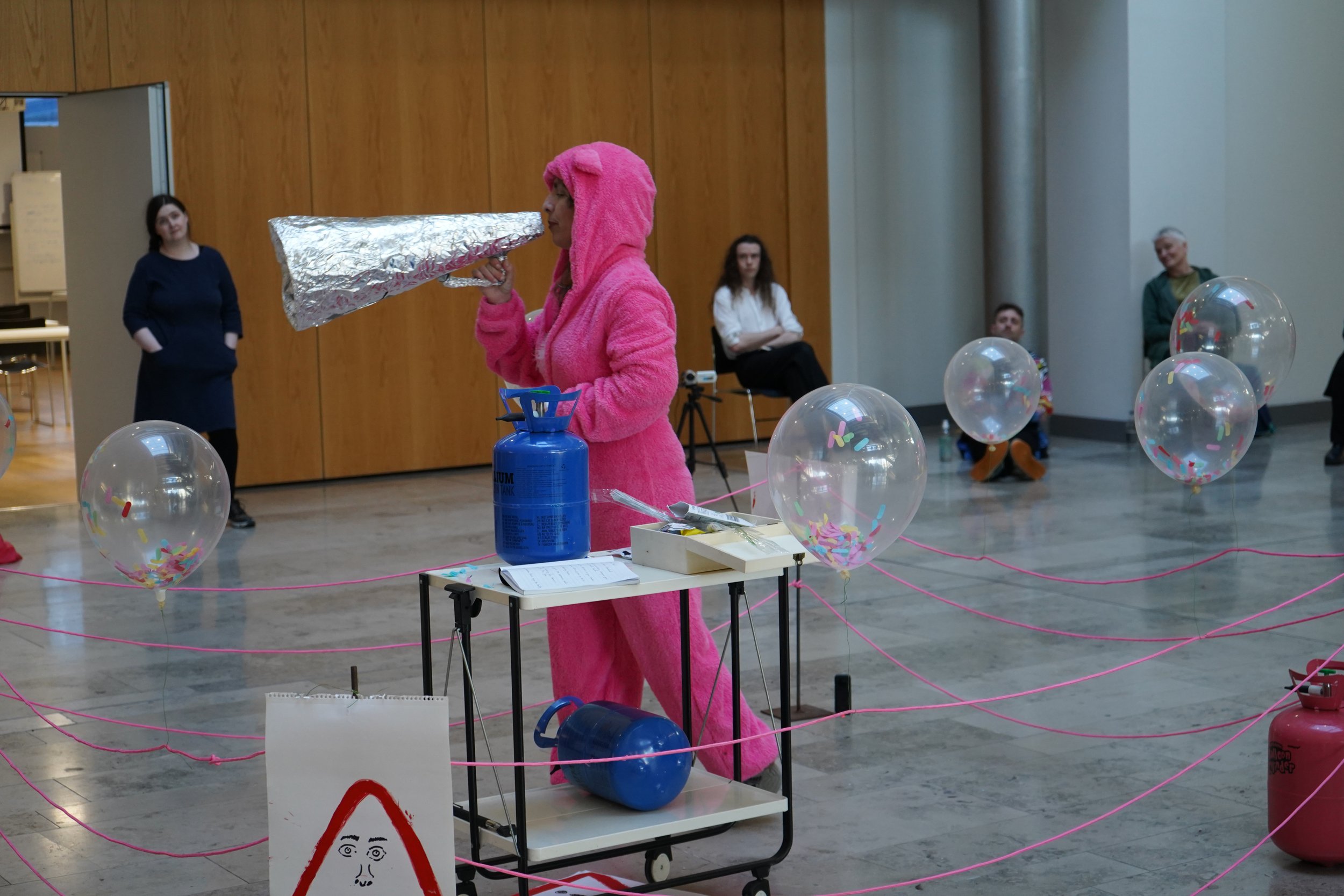 Person dressed in a pink fluffy costume with a hood resembling an animal, holding a large object wrapped in silver foil in a room with wooden walls and balloons, and observing a table with various objects and a sign.