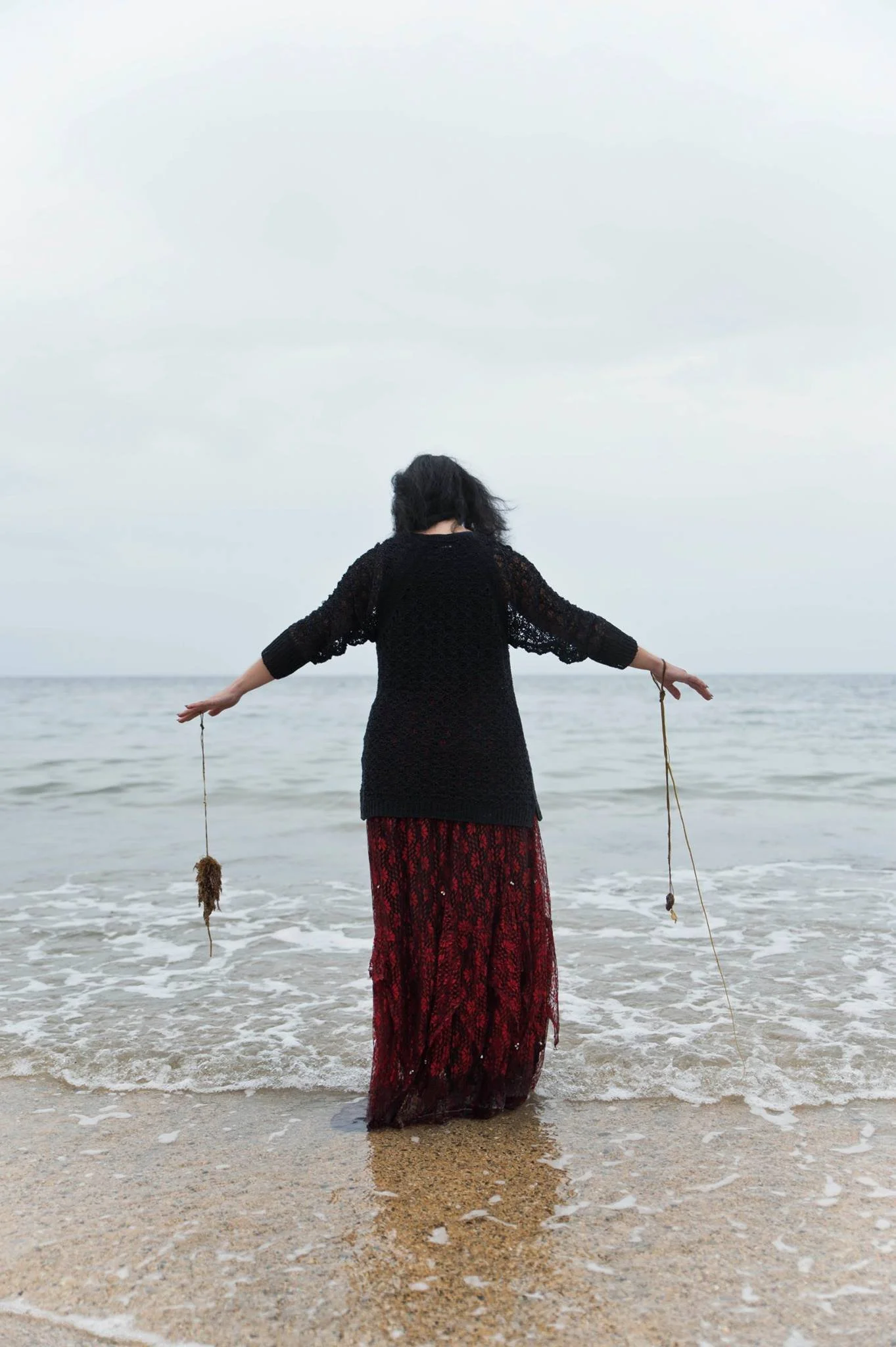 A woman wearing a black lace top and a long red patterned skirt stands in the ocean with her arms outstretched, facing the water and cloudy sky.