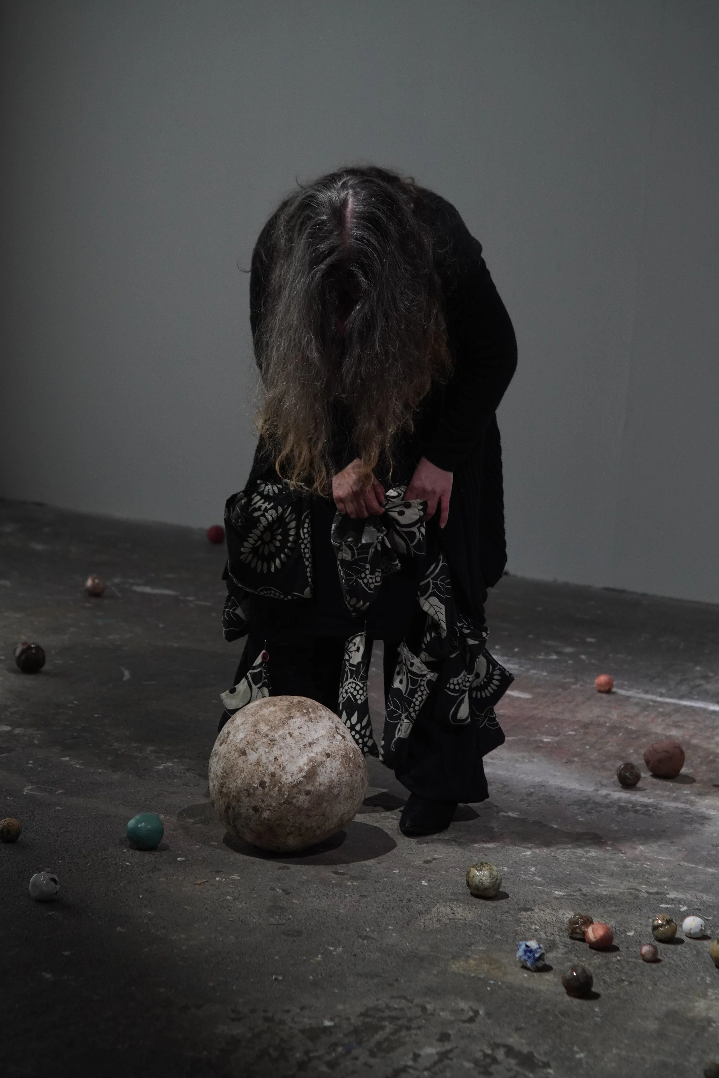 A person with long gray and brown hair, dressed in black, bending down, holding a black and white patterned cloth, surrounded by scattered balls and a large textured ball on a dark, worn floor against a plain gray wall.