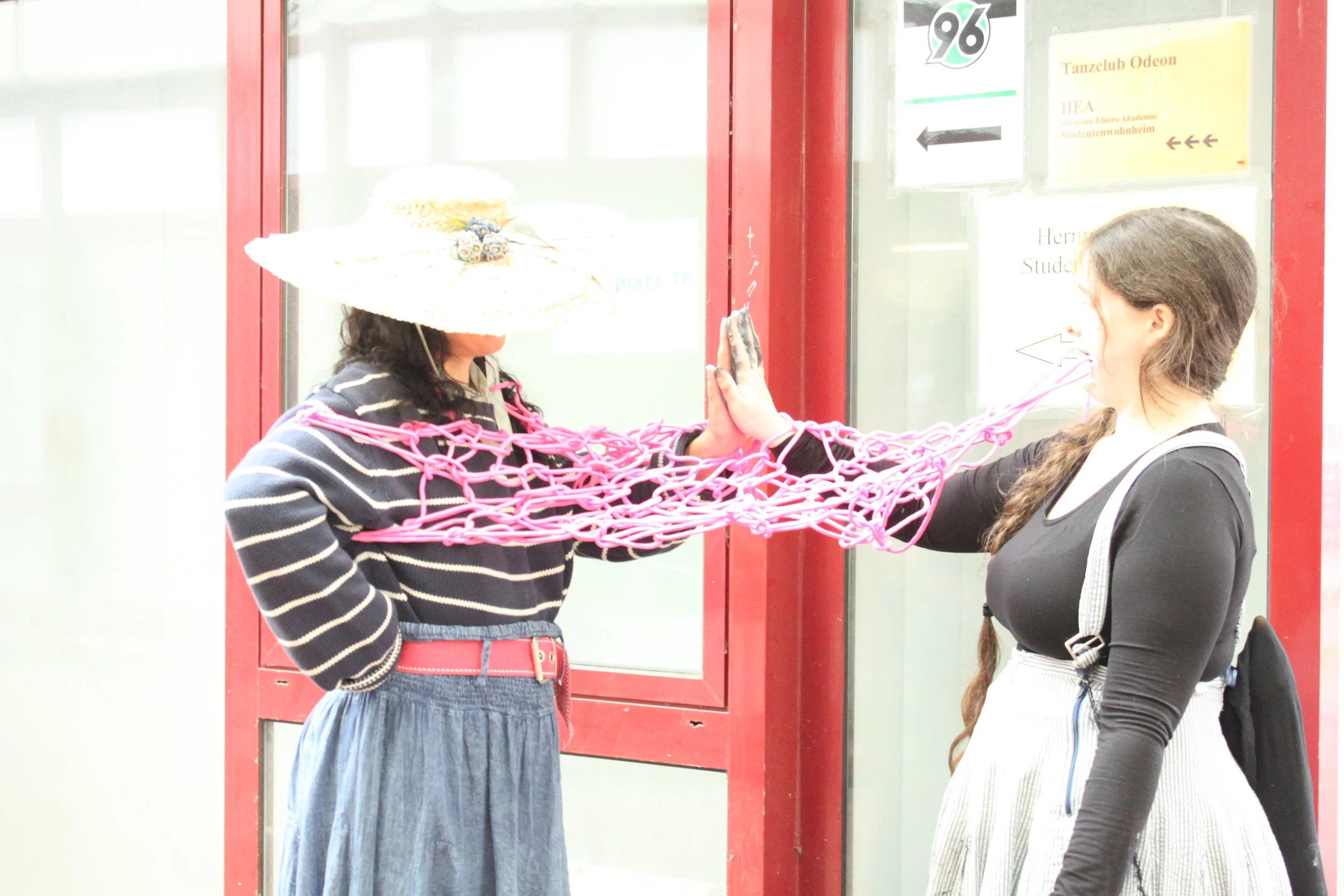 Two women are touching hands in front of a glass window behind which some signs and information are visible. One woman is wearing a striped sweater and a wide-brimmed decorated hat, the other woman has long braided hair and is wearing a black top wit