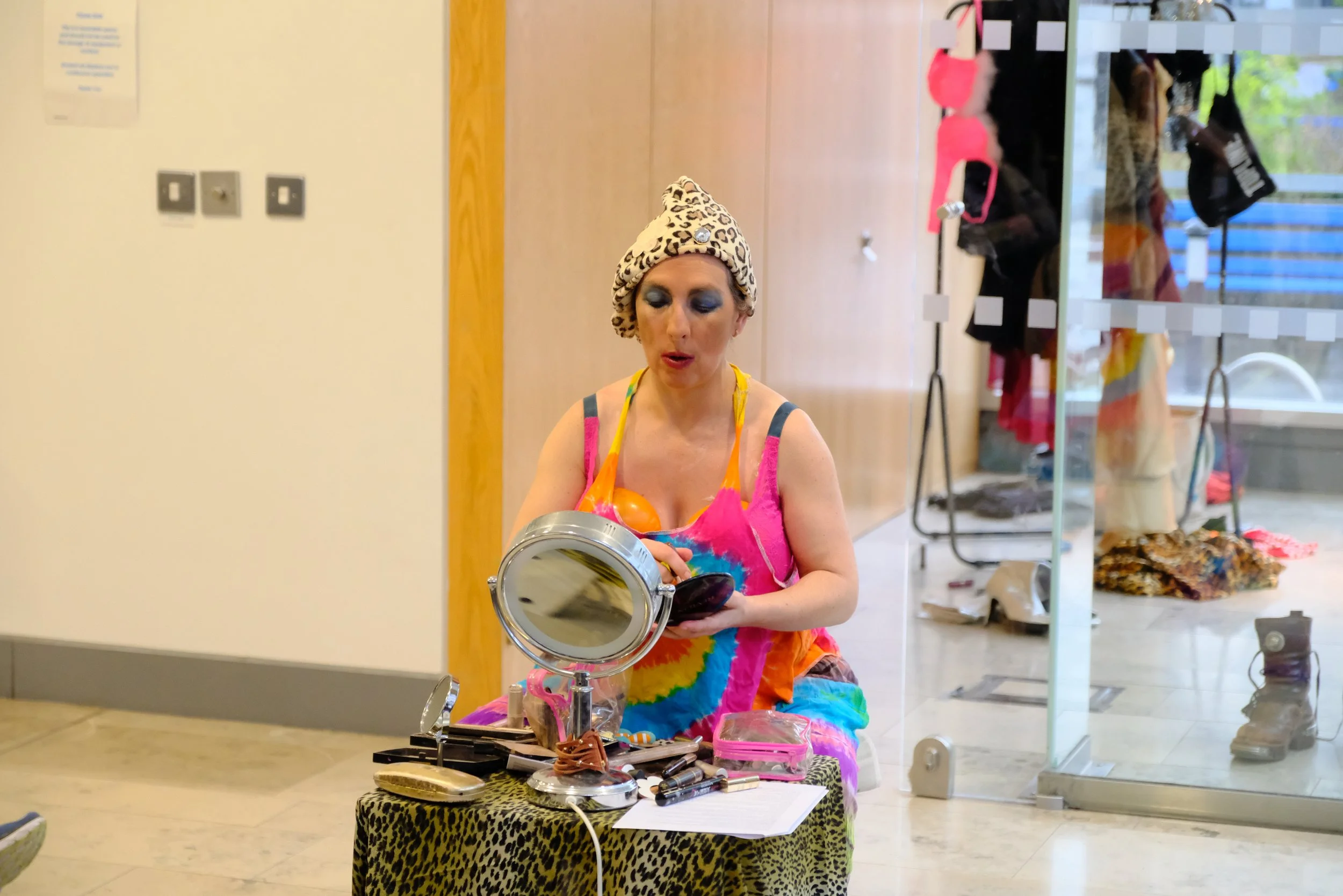 A woman with a leopard print headband and colorful tie-dye dress applying makeup at a mirror, surrounded by makeup products on a leopard print stool.