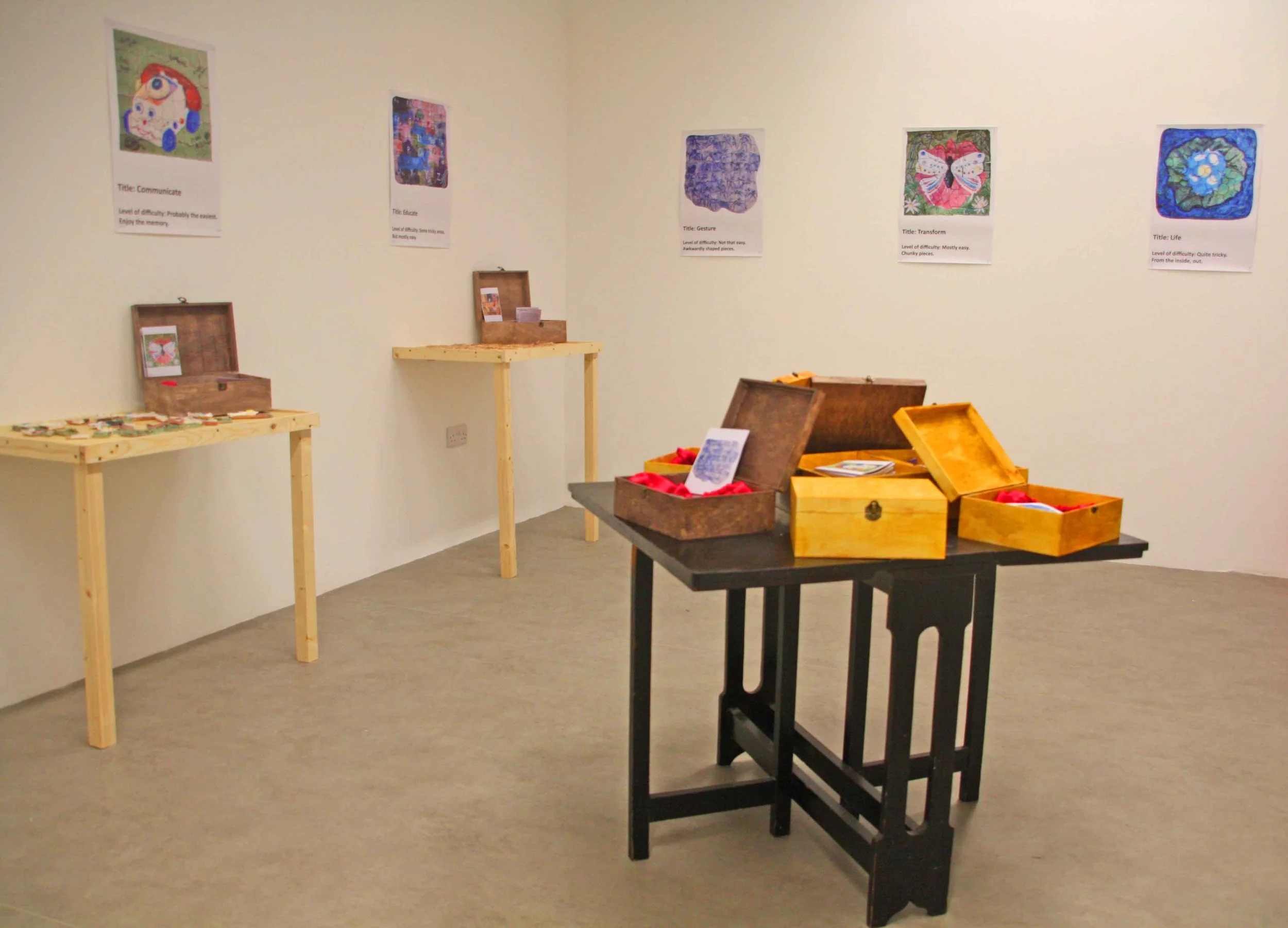 An art exhibit room with framed artwork on the wall, wooden display tables, and open wooden boxes with red and yellow cloth inside.