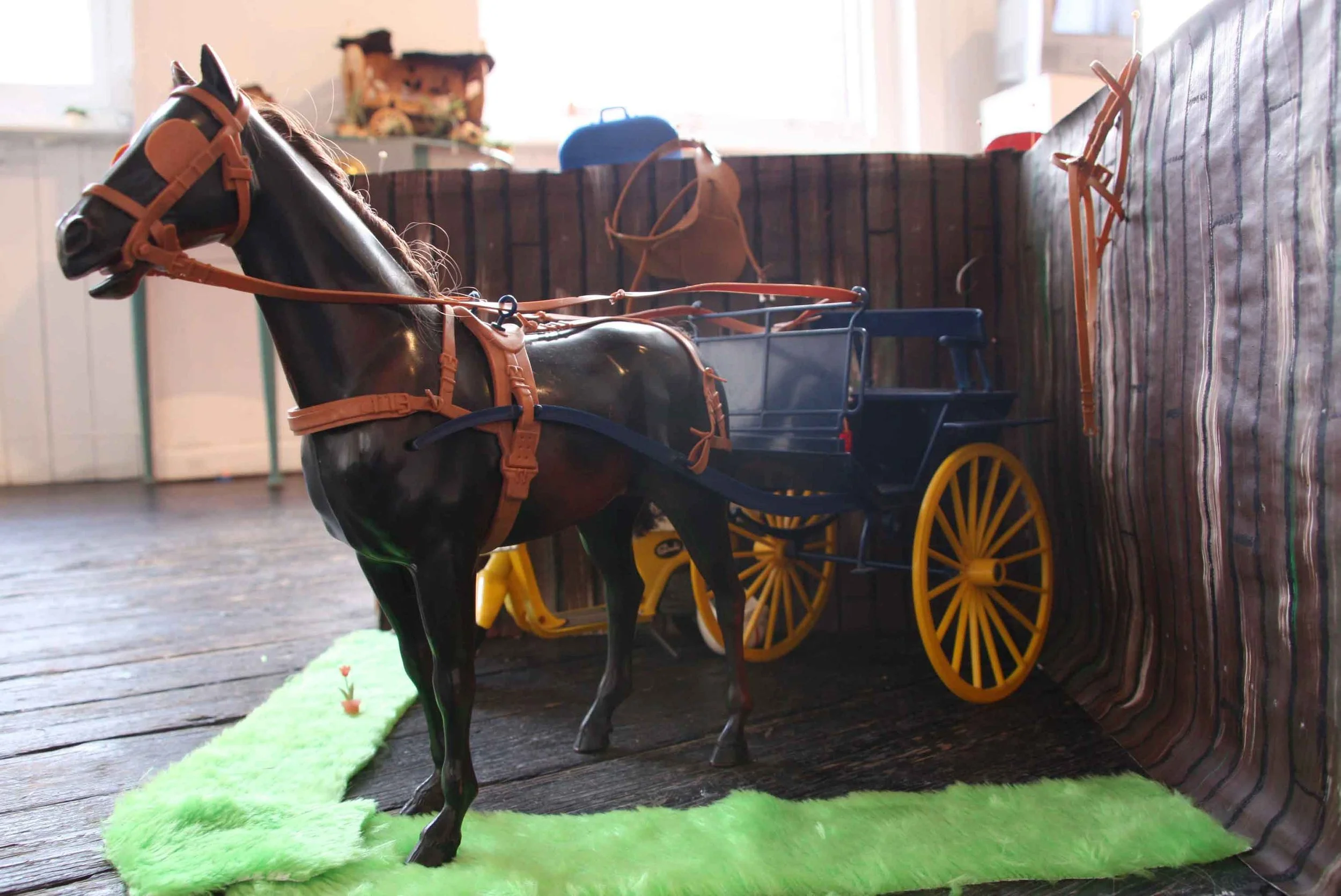 A toy horse attached to a small black and yellow cart. The toy horse is placed on a green felt mat on a wooden floor with a blurred background of a room with sunlight coming through the window.