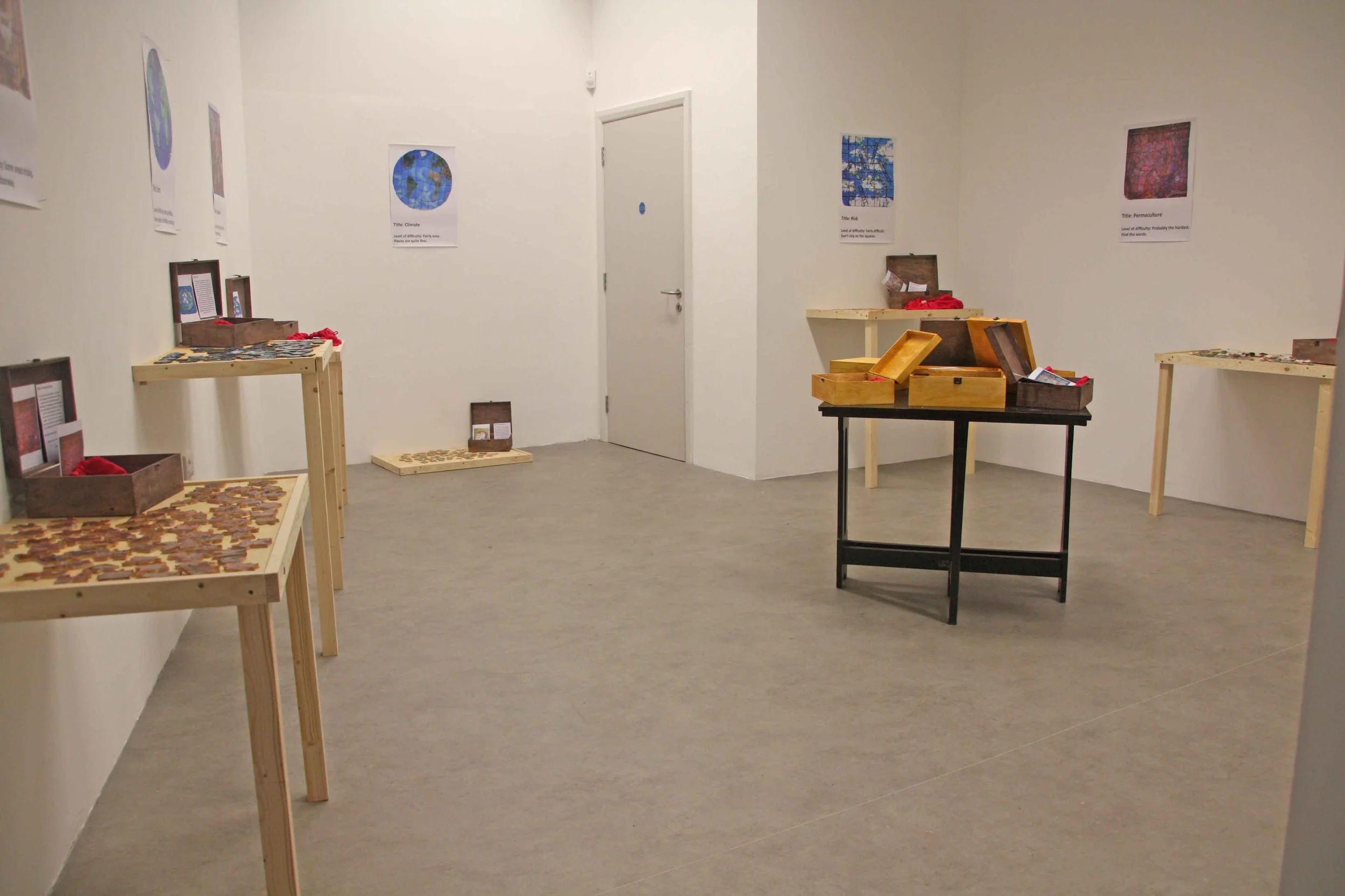An art exhibition room with white walls and light grey flooring, displaying posters of Earth with different themes such as climate and permaculture, and various wooden tables and boxes holding puzzle pieces and informational materials.