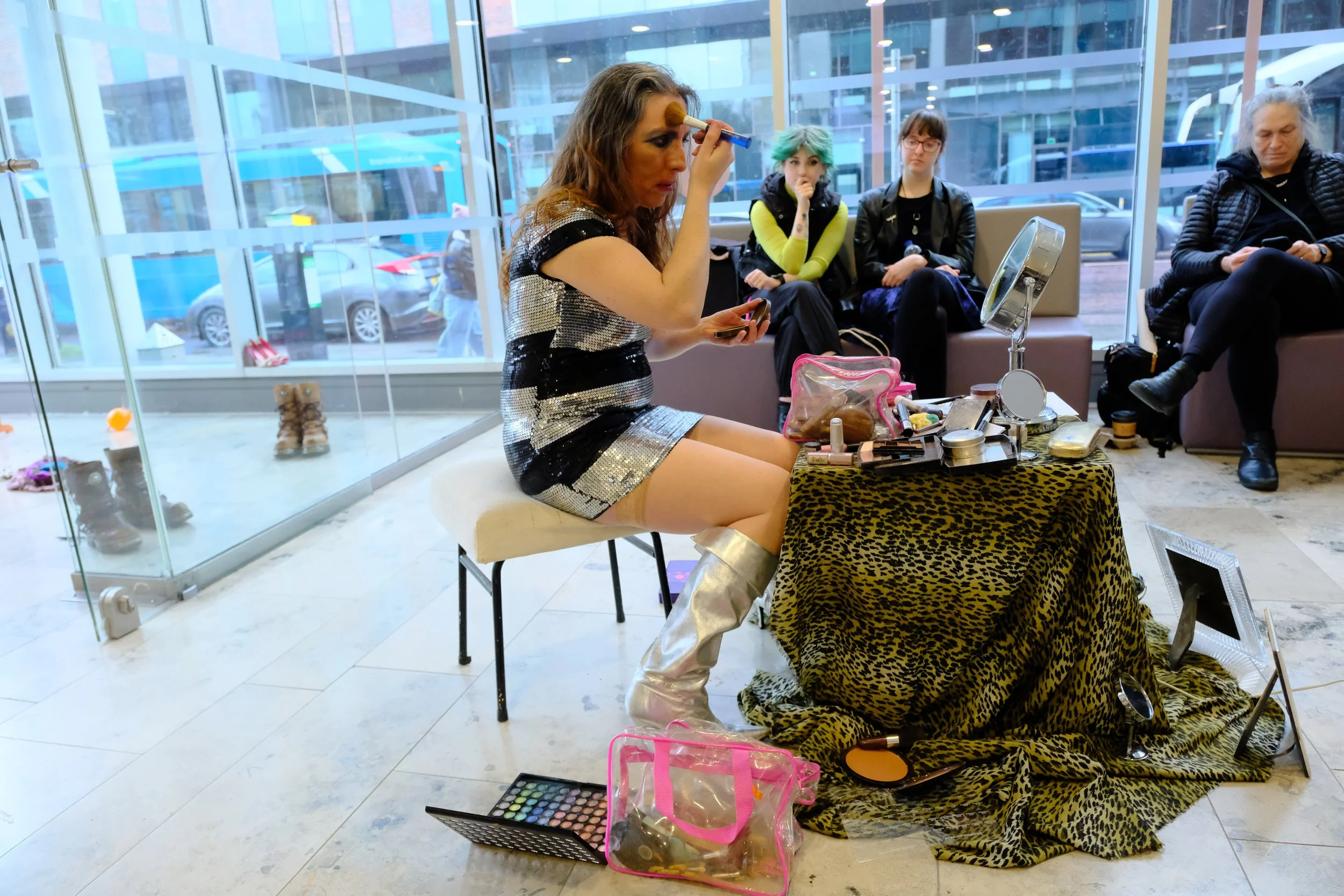 A woman sitting on a stool applying makeup to her face in a shopping mall, with three people seated on a bench behind her. The woman is dressed in a silver and black striped dress, has makeup brushes in her hand and makeup products on a table covered