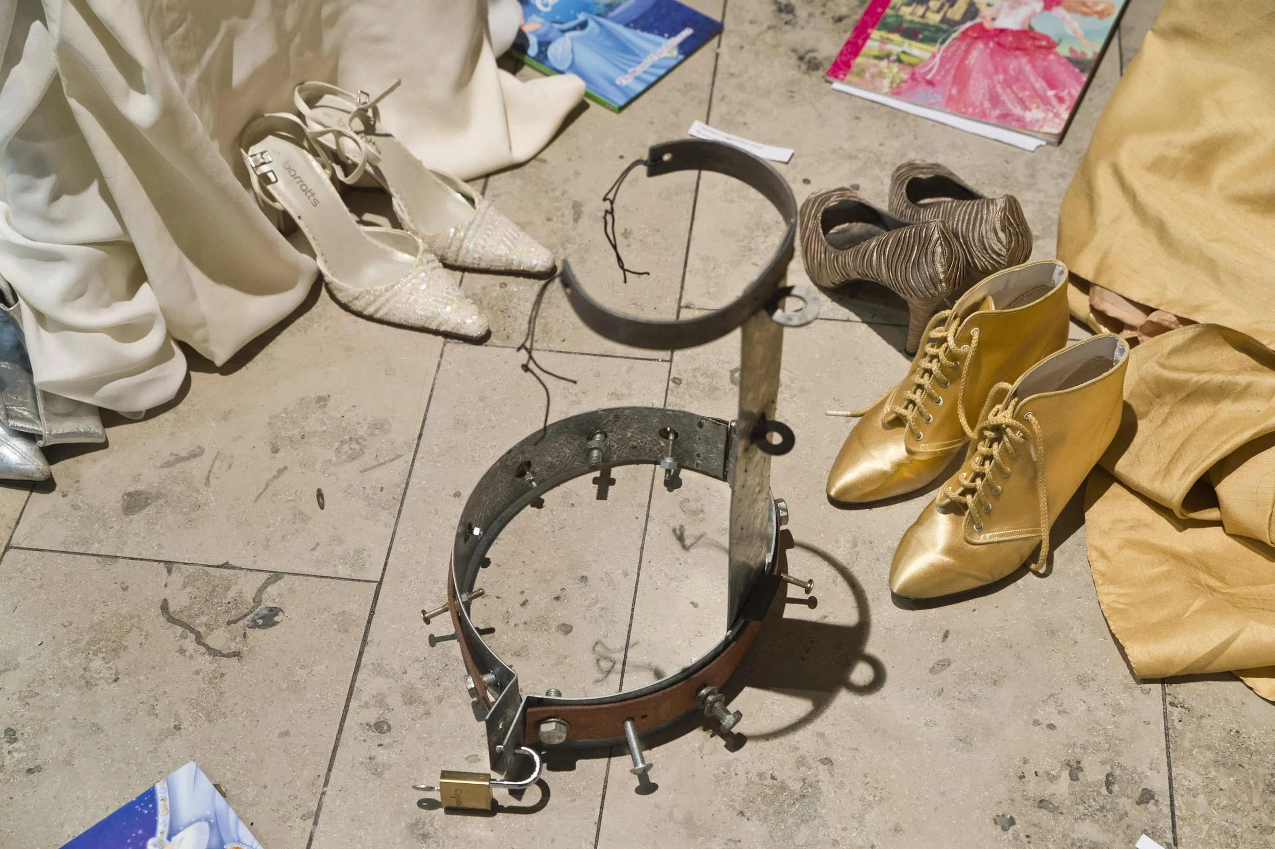 A collection of shoes on a concrete floor, including white glittery high heels, yellow combat boots, gray textured high heels, and beige shoes. There are also magazines and a black metal jewelry stand nearby.