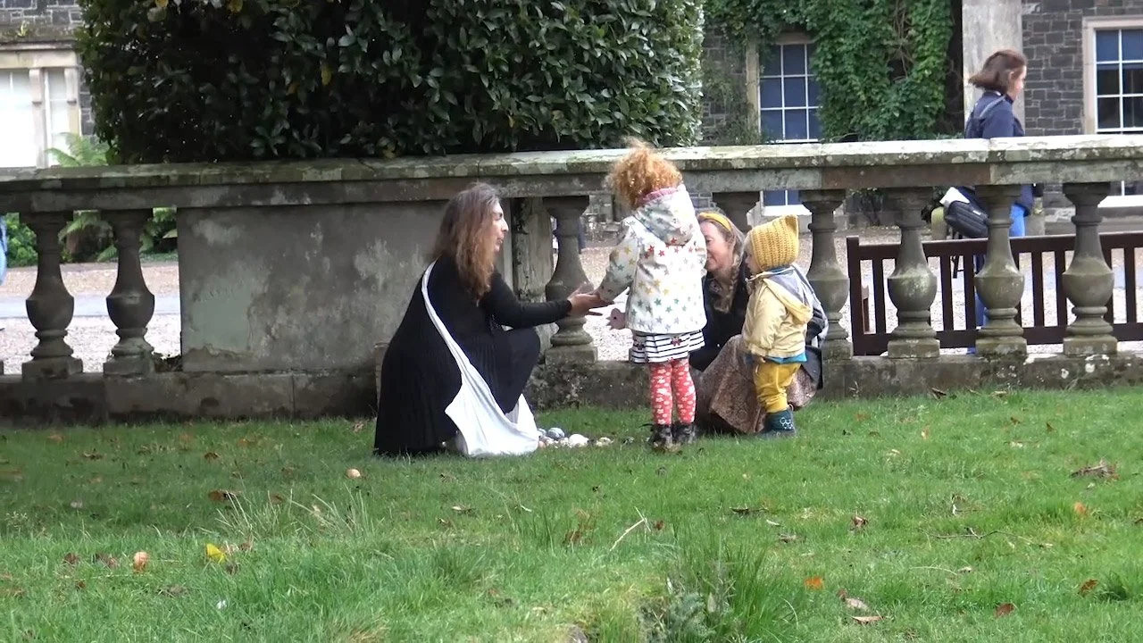 A woman kneeling on the grass, talking to two small children with an older woman nearby. The scene is outdoors next to a stone wall and a bench.