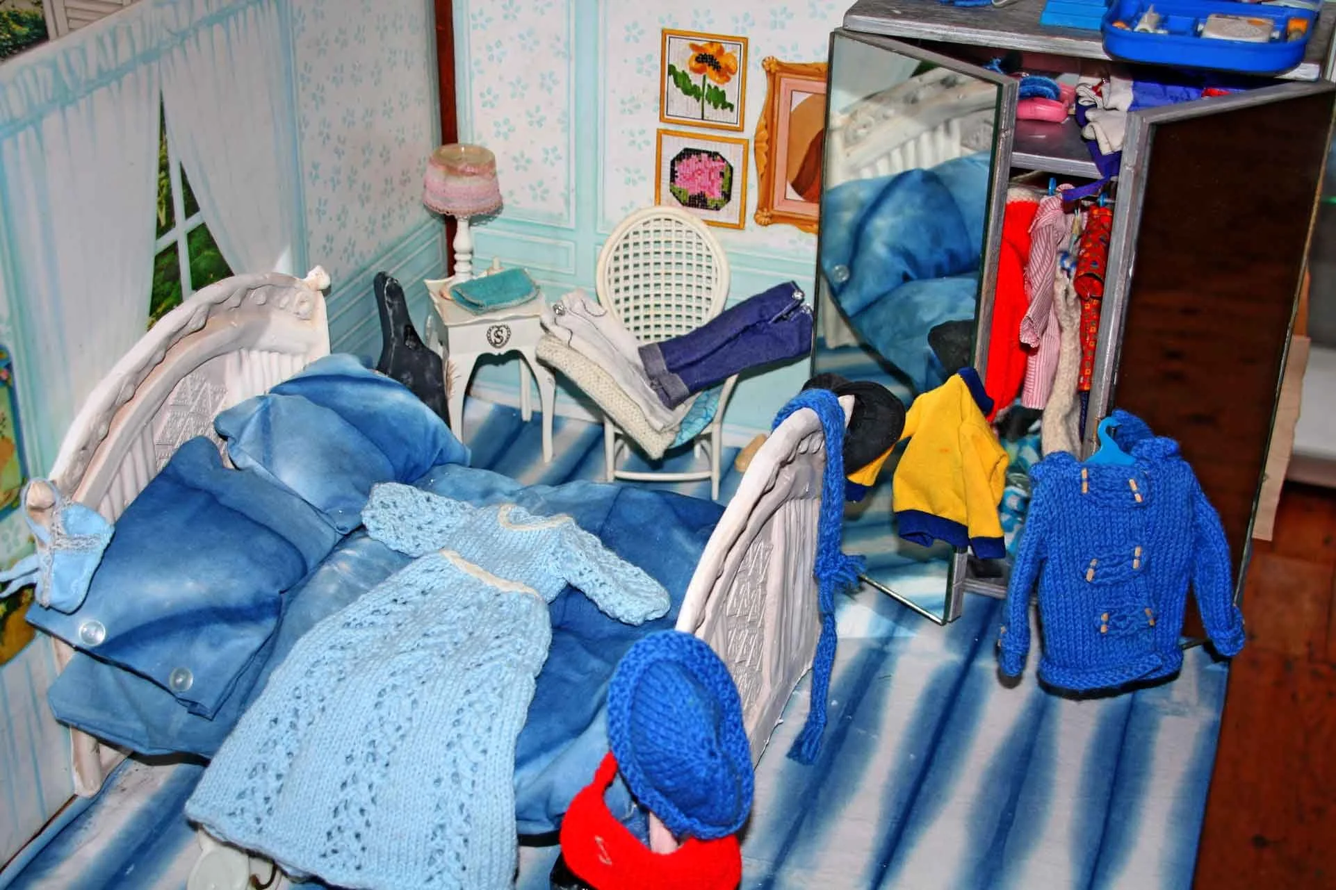 Cluttered doll house bedroom with a blue bed, pile of clothes, a small white table with a folded towel, an elaborate headboard, a wicker chair, wall artwork, a mirror, and a dresser with drawers open and clothes spilling out.