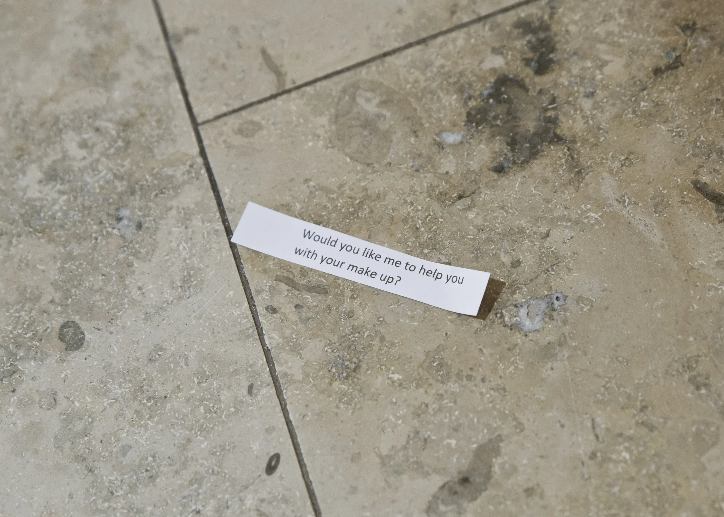 Close-up of a tiled floor with a white piece of paper that has text on it. The paper reads: 'Would you like me to help you with your make up?'