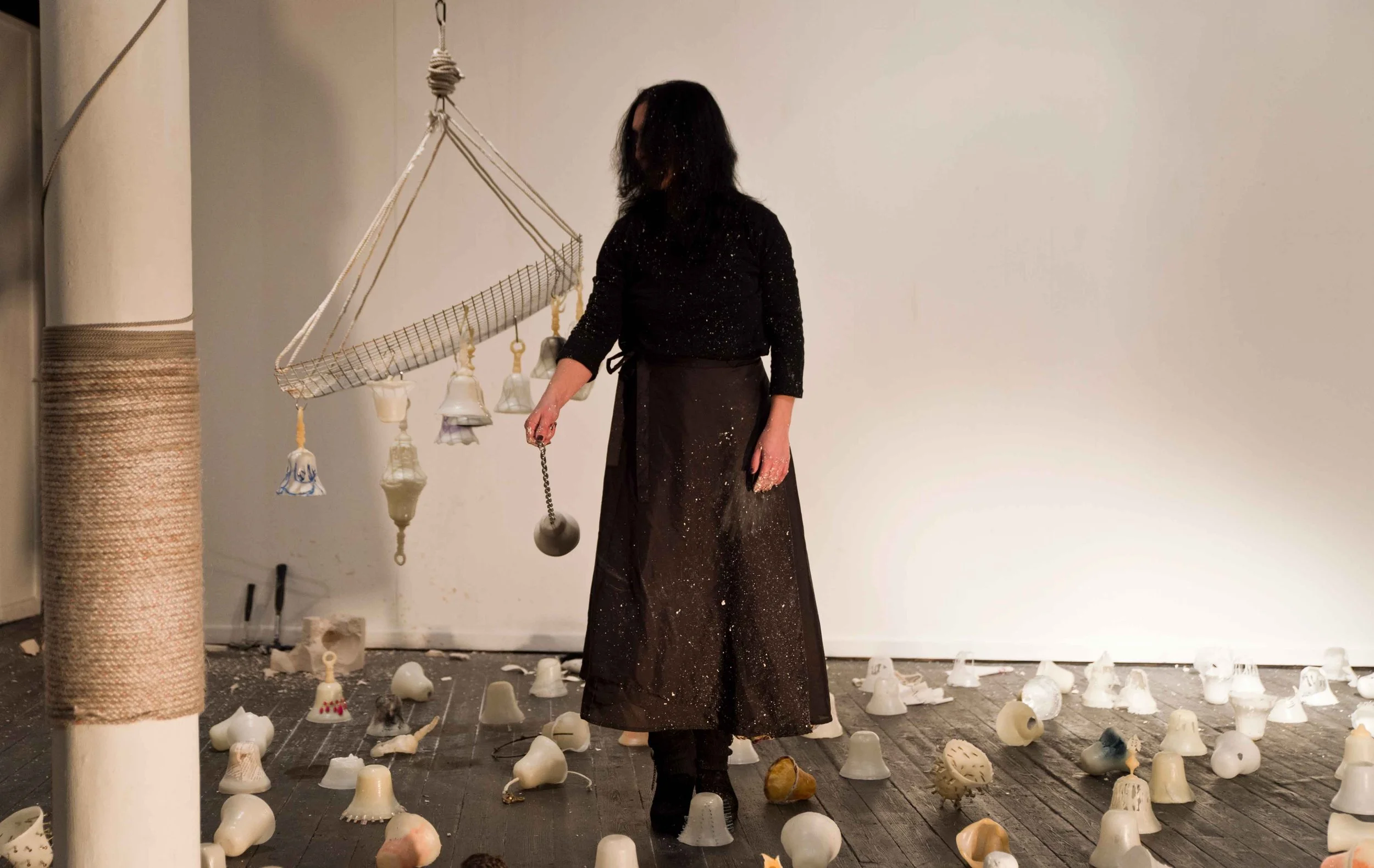 A woman standing in an art installation room, surrounded by broken or discarded ceramic objects on the floor. She is facing away from the camera, wearing a long black skirt and top with glitter. Behind her is a wall with a hanging mobile with bells o