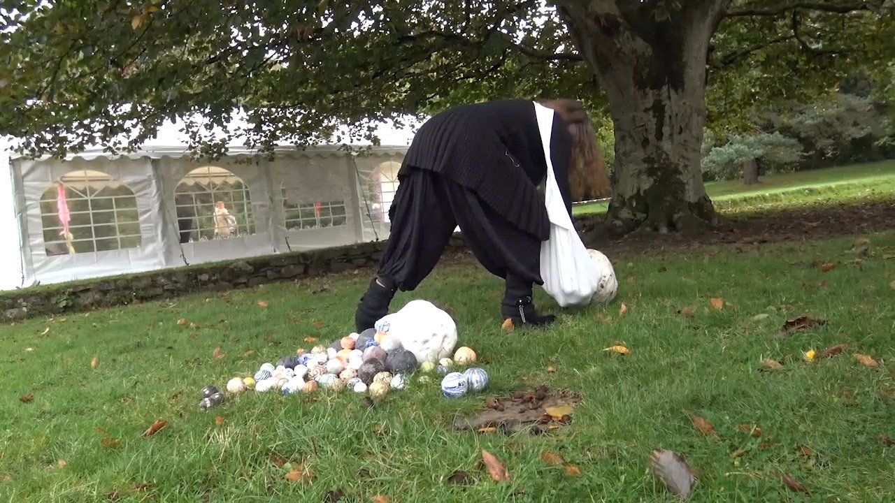 A person dressed in dark, medieval-looking clothing is bent over, scraping a large skull on the ground. They are near a pile of golf balls. Behind them is a large tree and a white tent with windows.