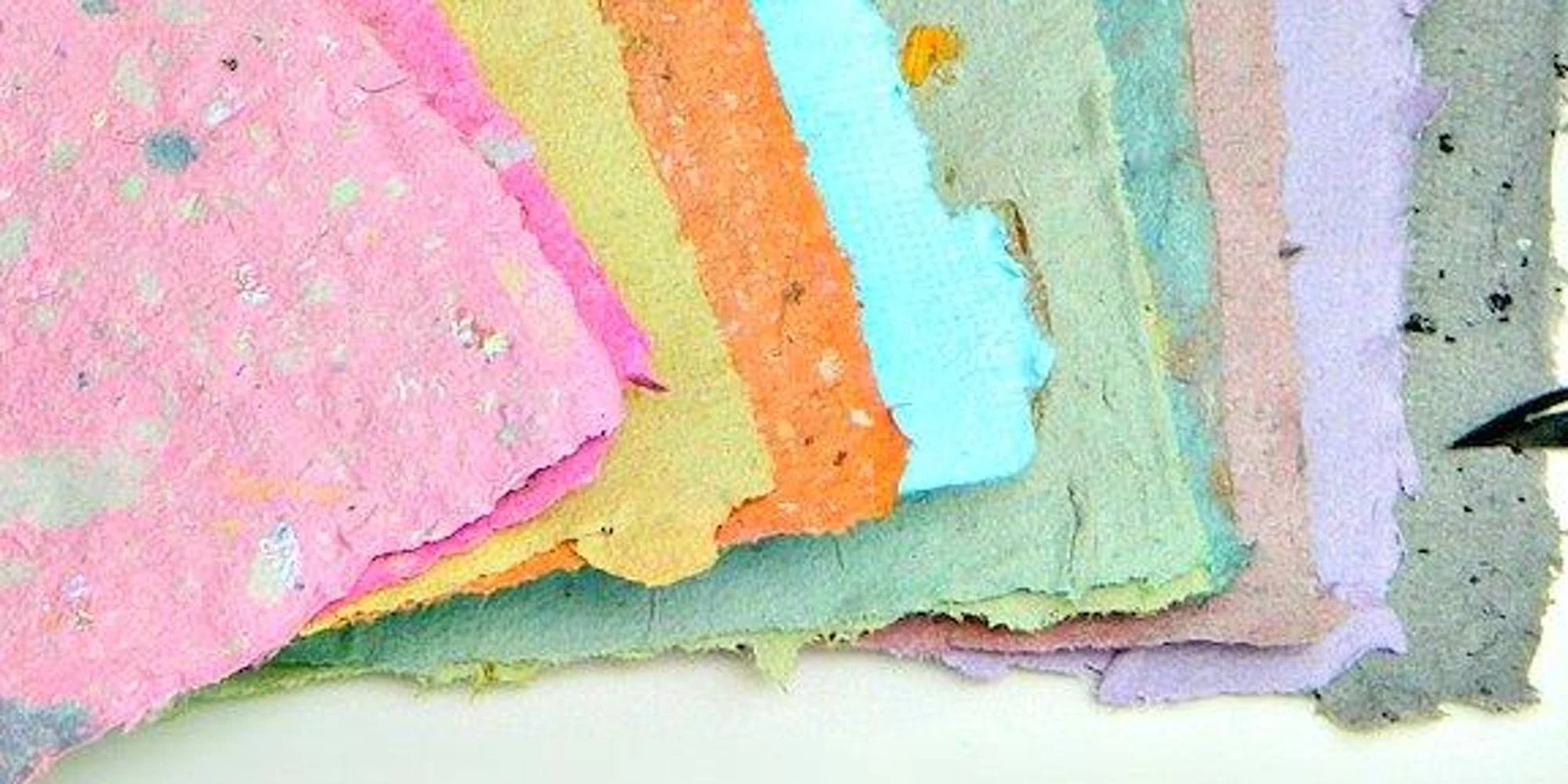 Close-up of layered pastel-colored watercolor paper in shades of pink, yellow, orange, green, blue, and purple, with some rough and torn edges.