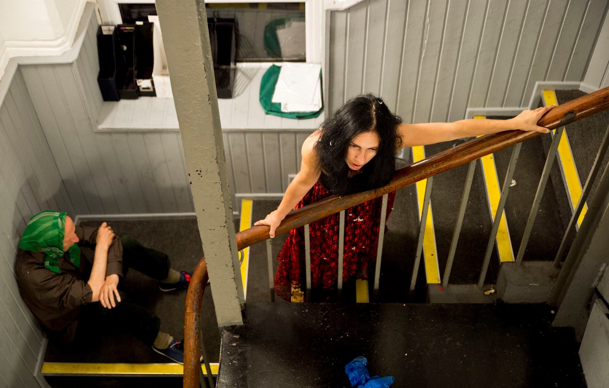 Woman in a black and red floral dress climbing a staircase with a wooden handrail in a small hallway. A person in a green hoodie with a hood is sitting on the floor near the wall, holding a phone, while a person with dark hair, dressed in black, look