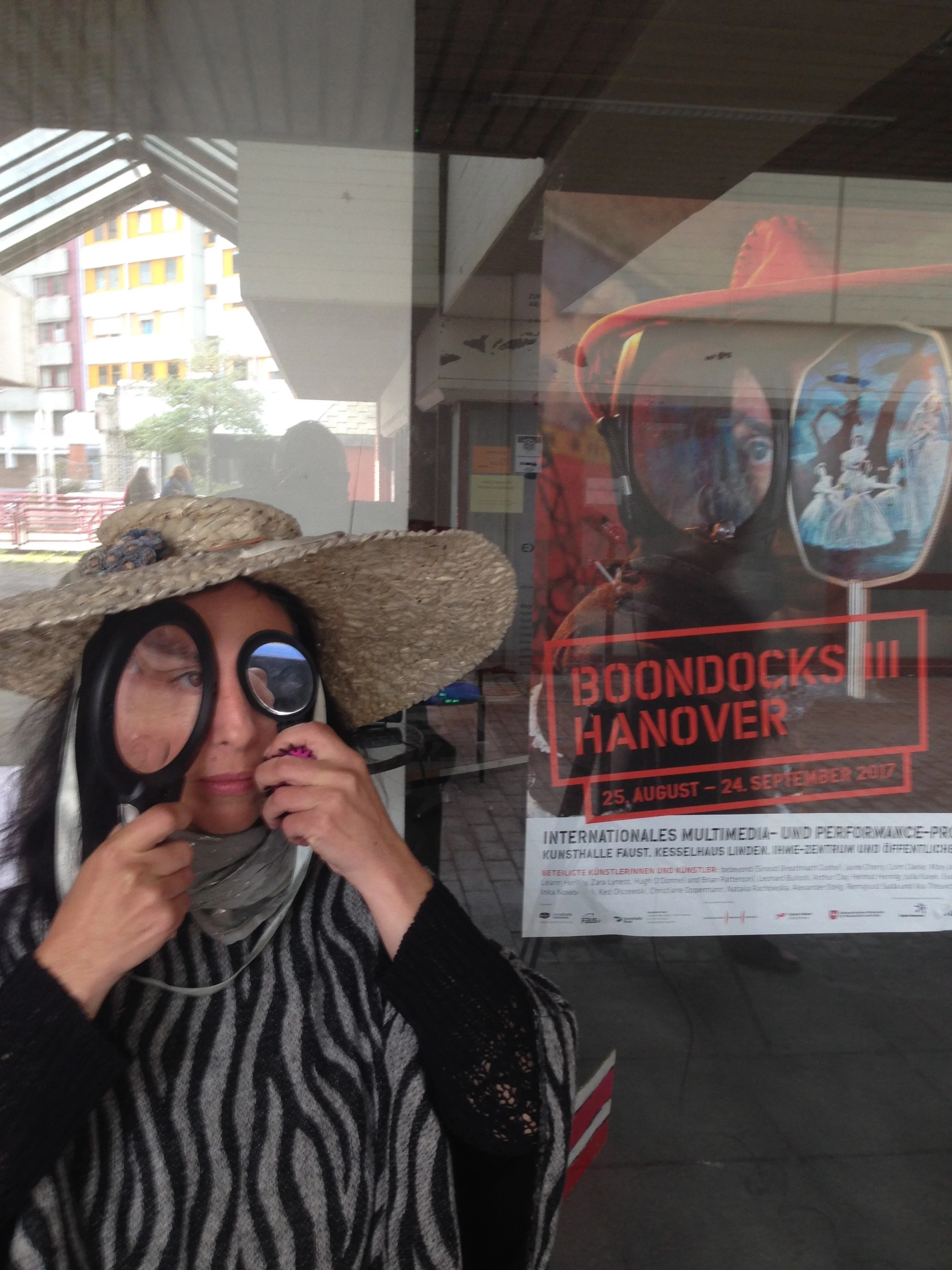 A woman with long gray hair, wearing a large straw hat with a decoration, glasses, and a black-and-white striped sweater, is holding binoculars and looking through them.