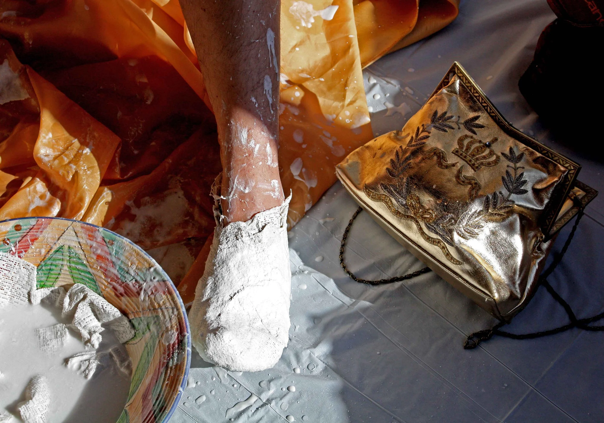 Close-up of an art or craft project on a table, including a decorative plate, a piece of cloth with a gold and black design, and a surface covered with a thick white substance, possibly glue or paint, with some splatters.