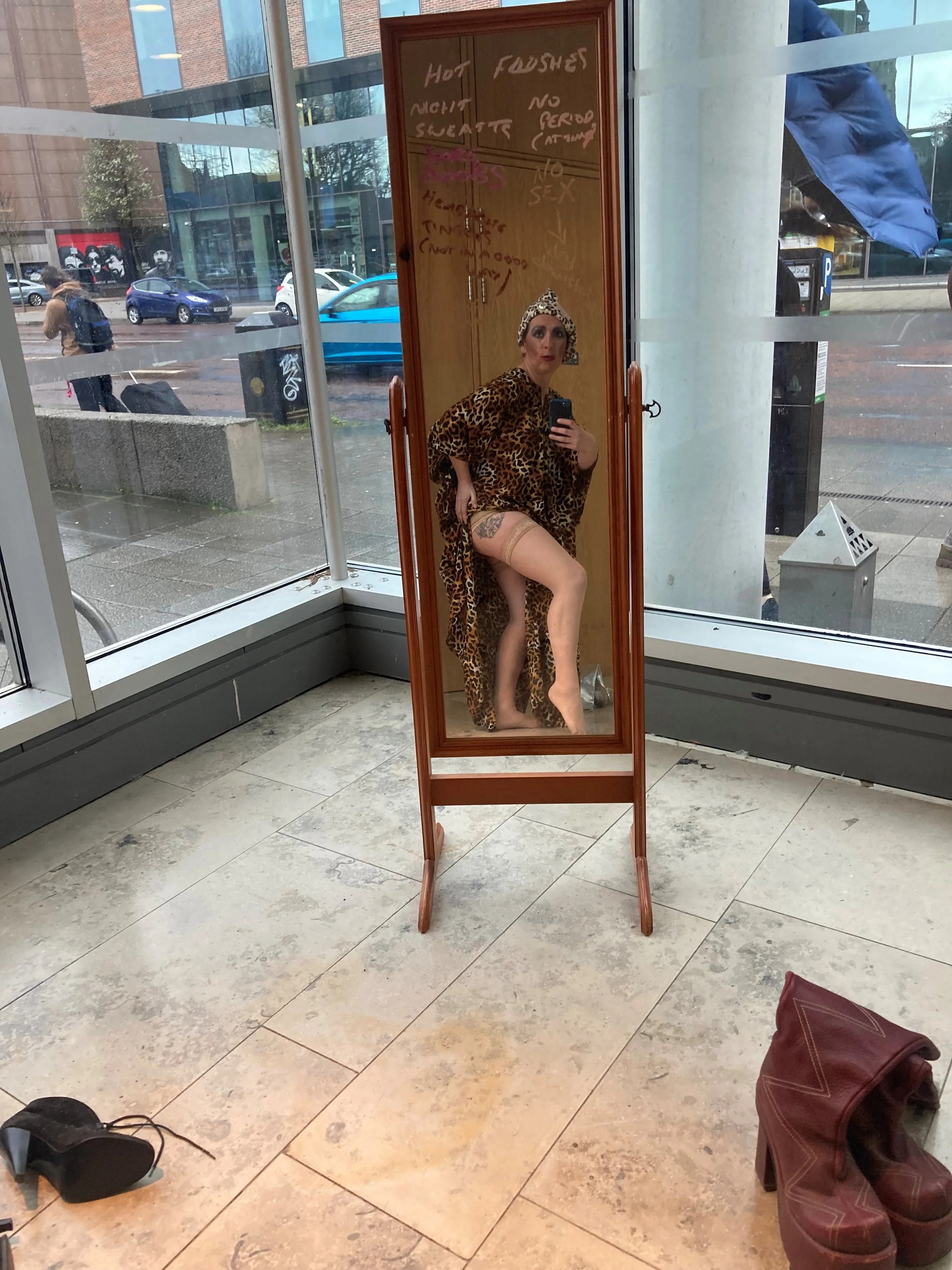 Woman in leopard print dress and headwrap taking a selfie in a full-length mirror inside a building with glass walls and outside street view.