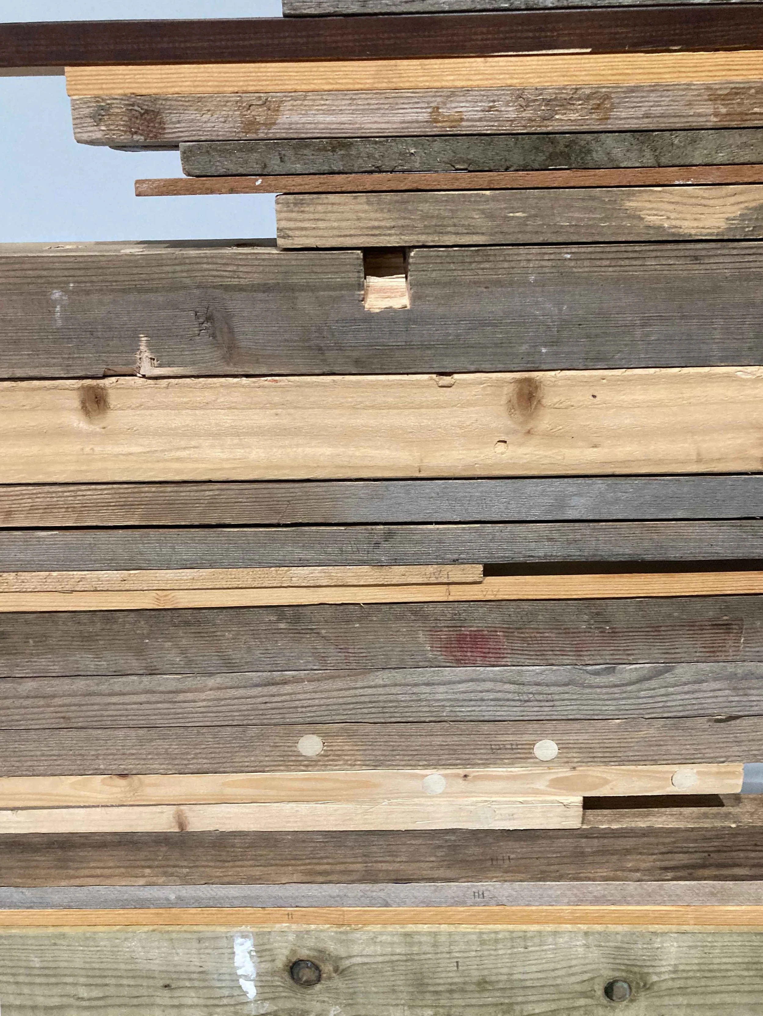 Close-up of stacked wooden planks of various colors and finishes, some with visible knots, nails, and weathered surfaces.